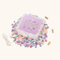 Stay Golden Bead Kit Rylee Ta with purple box lid and loose letter beads, pastel star and heart beads and elastic cord spilling out, flat lay on cream background