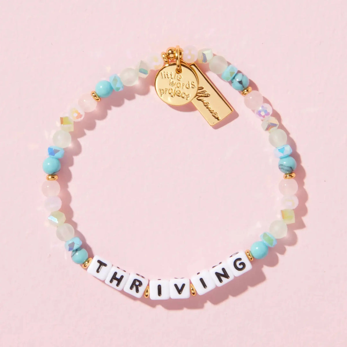Thriving bracelet by Katie Thurston, white letter beads with turquoise stone, pink, sea green frosted glass, and iridescent crystal beads with gold accents and gold Little Words Project and Kindfluencer tags, flat-lay on pink background