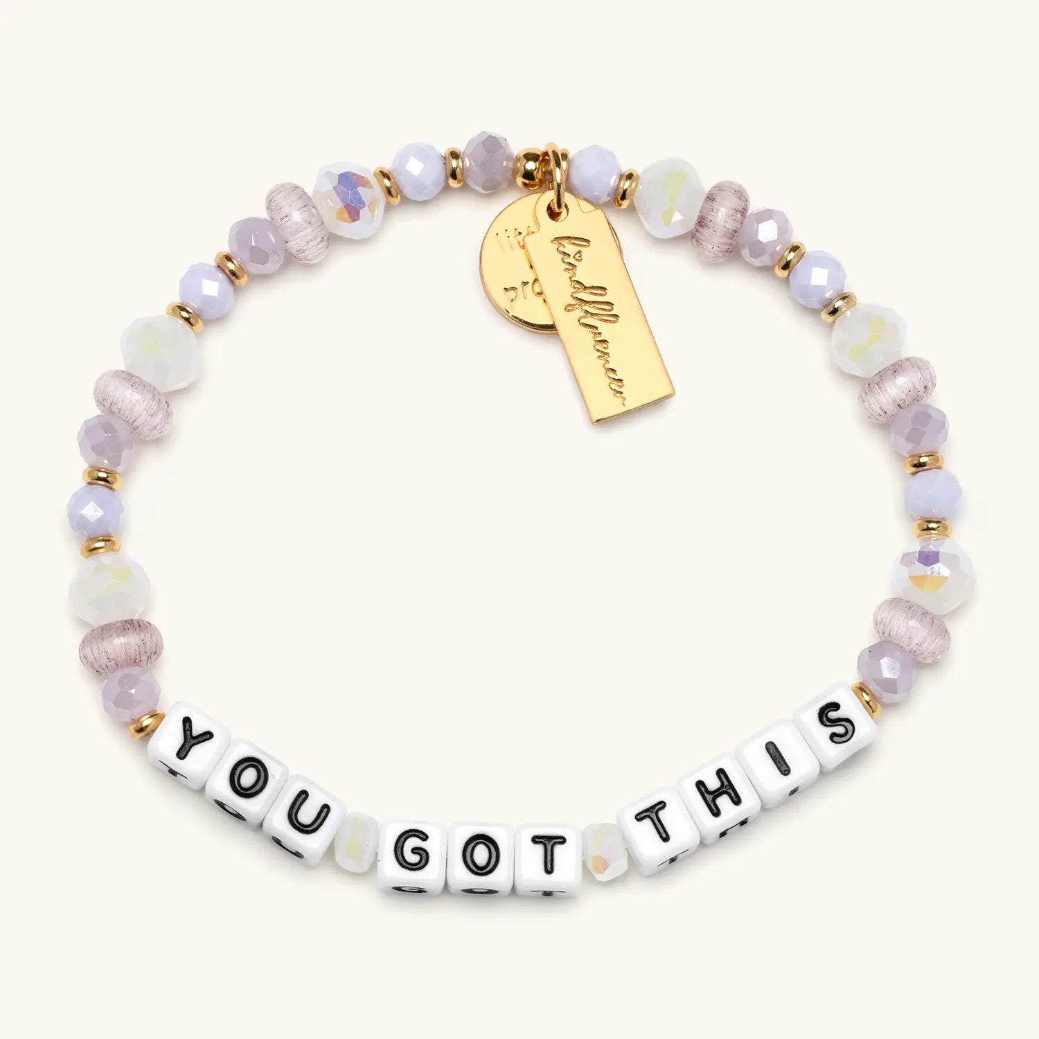 A single beaded bracelet spelling "YOU GOT THIS" in white square letter beads with black text. The bracelet features a mix of faceted lavender, lilac, and iridescent white beads with small gold spacers, giving it a soft, sparkly appearance. A gold rectangular charm with cursive branding hangs from the bracelet, displayed on a white background.