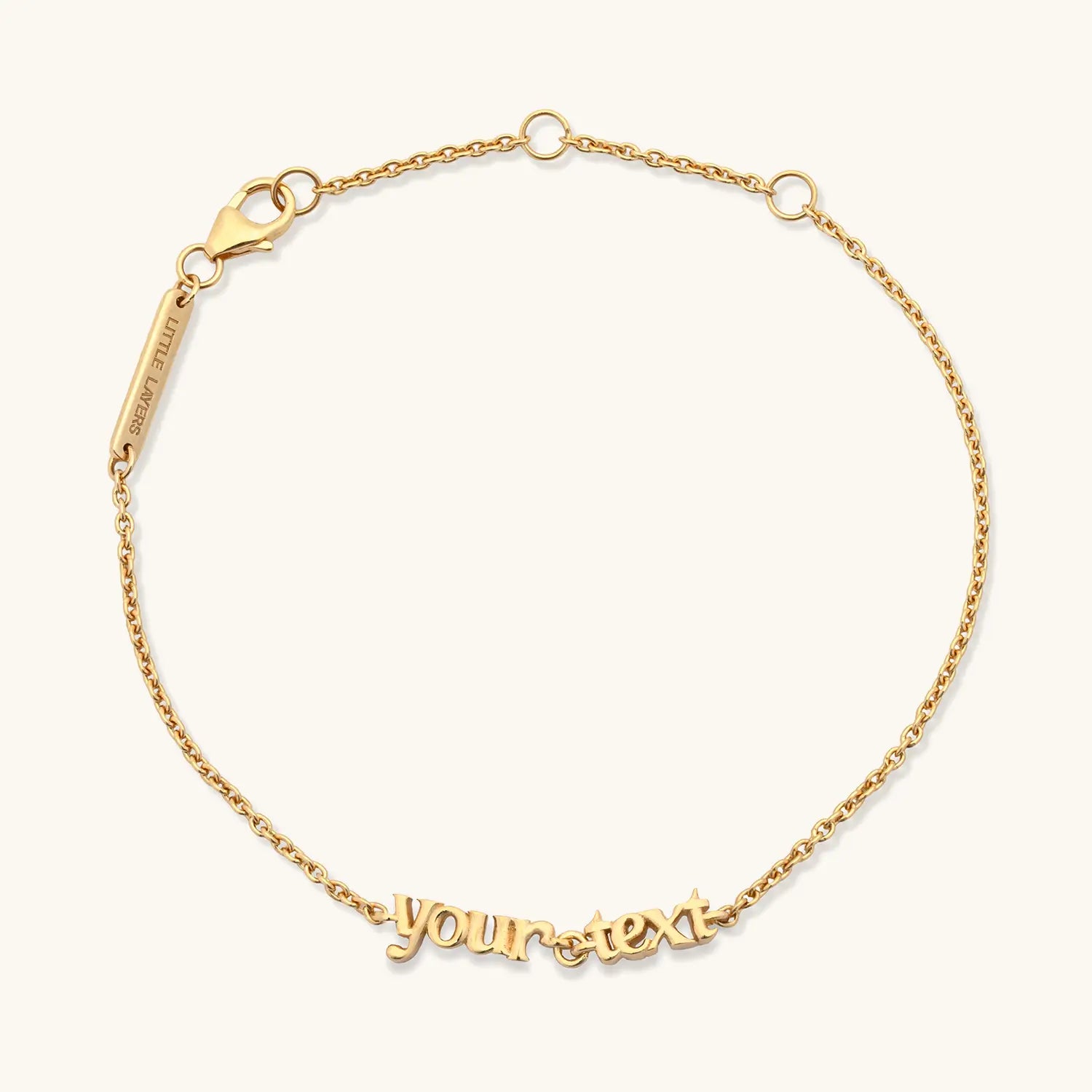 A delicate gold chain bracelet with a personalized script nameplate reading 'your text' and a lobster clasp, shown on a light cream background.