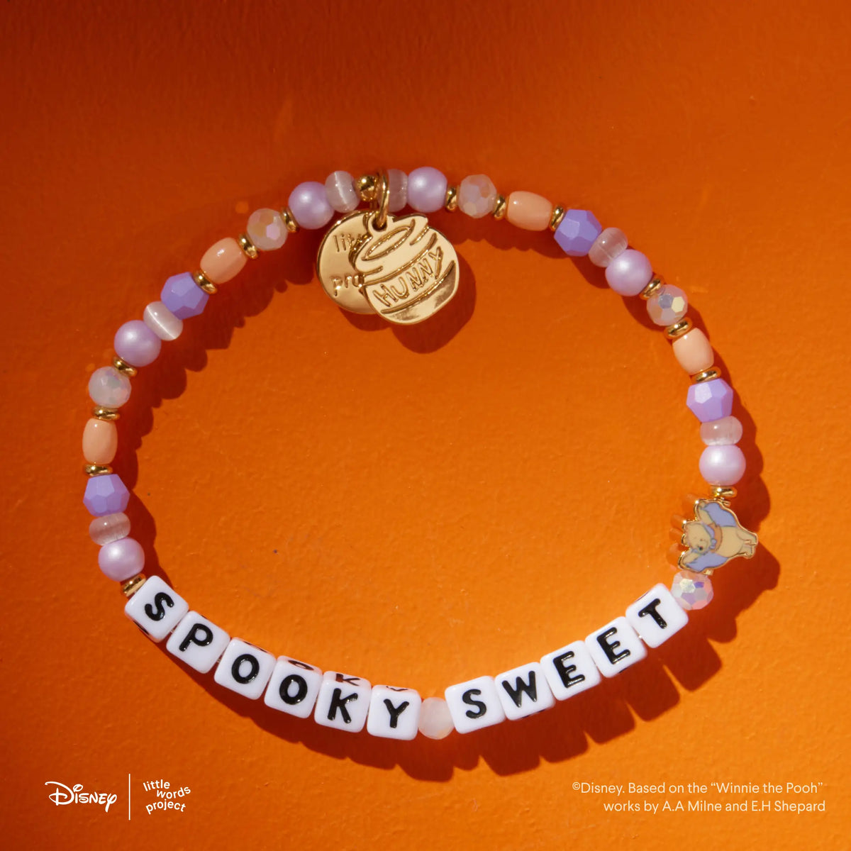 Spooky Sweet Disney's Winnie the Pooh bracelet in the Spooky Sweetie bead pattern, white letter beads with lavender, peach, and pearl beads, gold hunny pot charm and Winnie the Pooh vampire casted bead, flat-lay on orange background