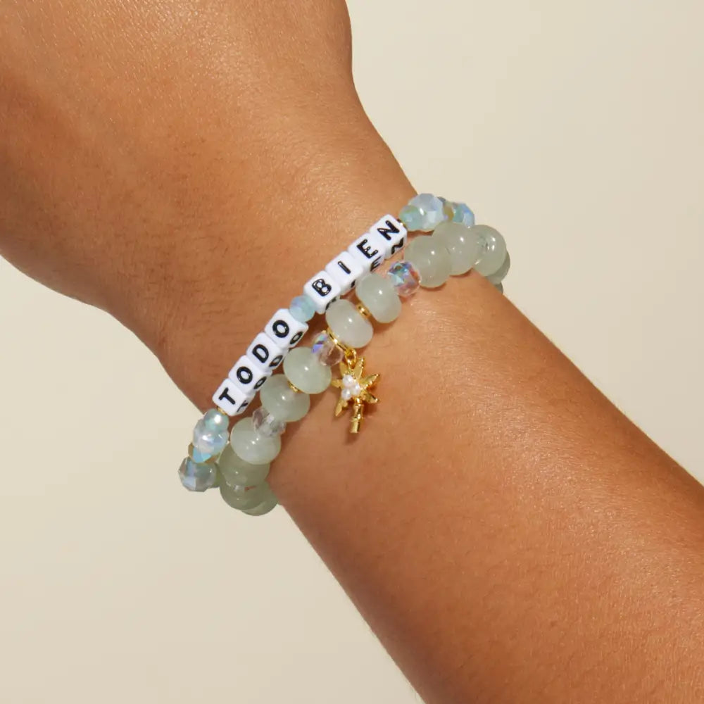 Todo Bien Bracelet Set by Kindfluencer Alexia Del Valle worn on wrist, white letter beads spelling Todo Bien on a Beach Glass pattern stacked with a Palm Kissed beaded bracelet and gold palm tree charm