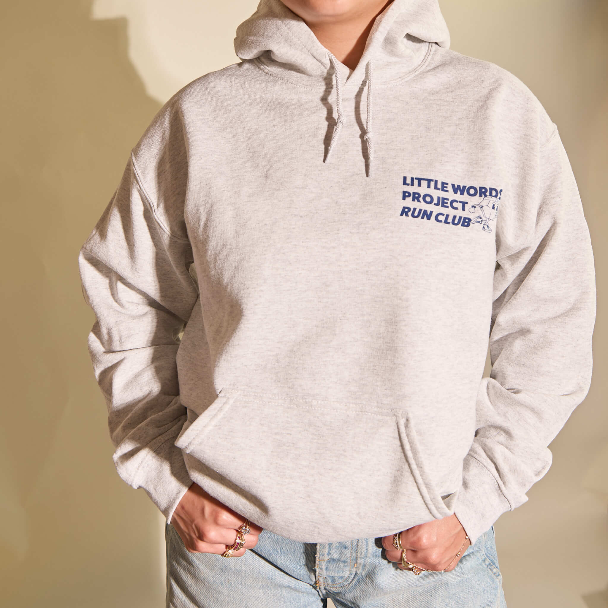 Person wearing a light gray hoodie with 'Little Words Project Run Club' text on a beige background