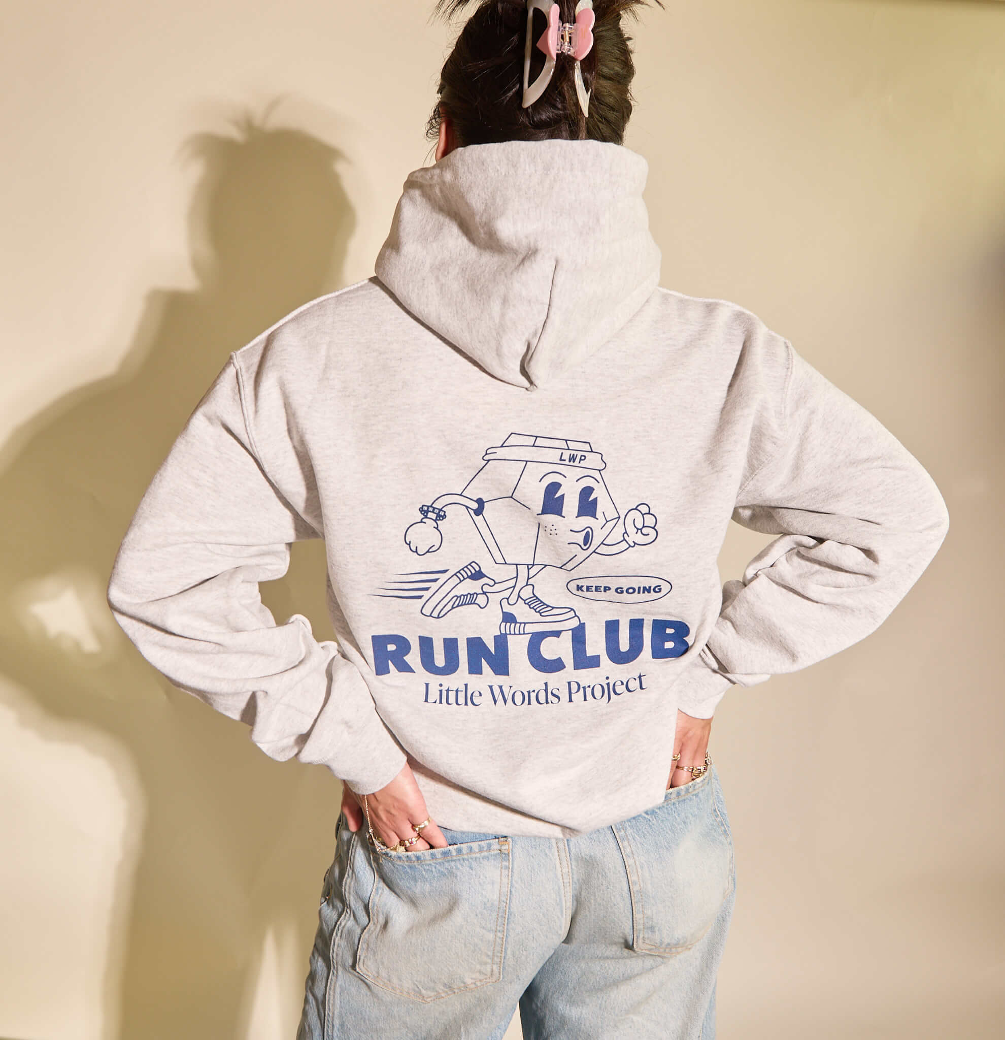 Person wearing a gray hoodie with 'Run Club Little Words Project' design on the back against a beige background