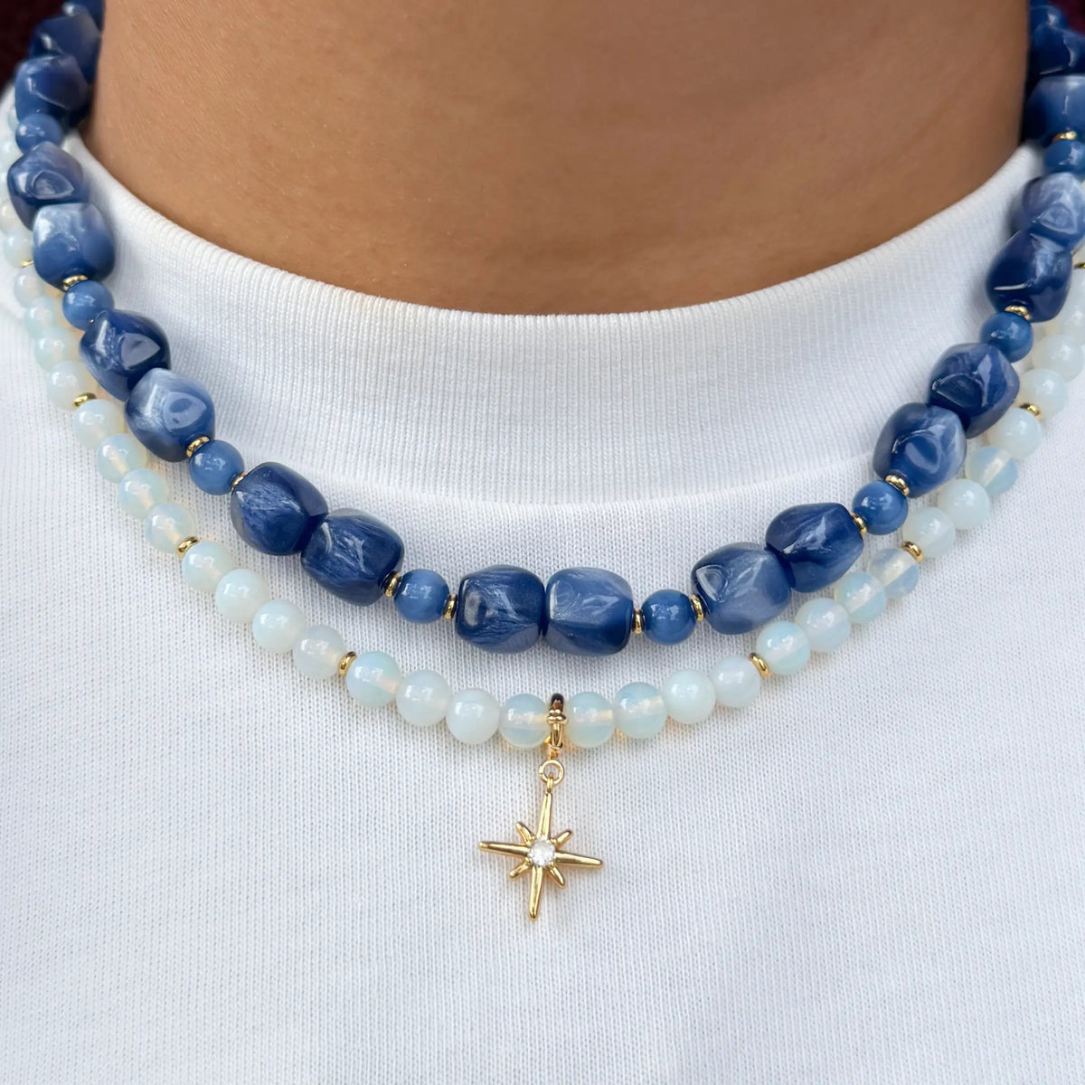 Radio Fever Charm Necklace Set and Opal Light Charm Necklace Set layered on-model, shimmery blue beaded necklace over a white opal beaded necklace with a gold starburst charm, worn with a white tee