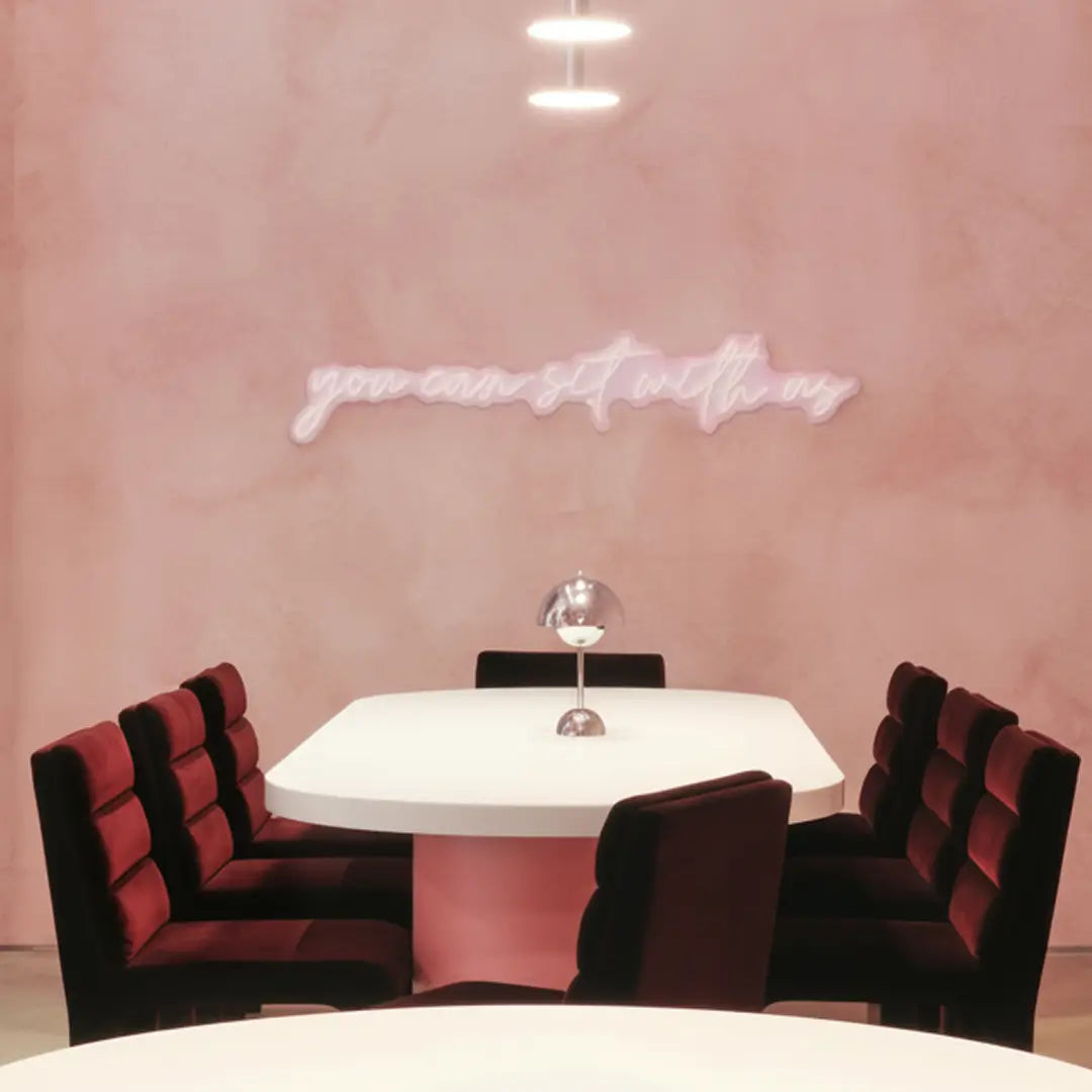 Modern dining area with red chairs and a white table against a pink wall with a neon sign.