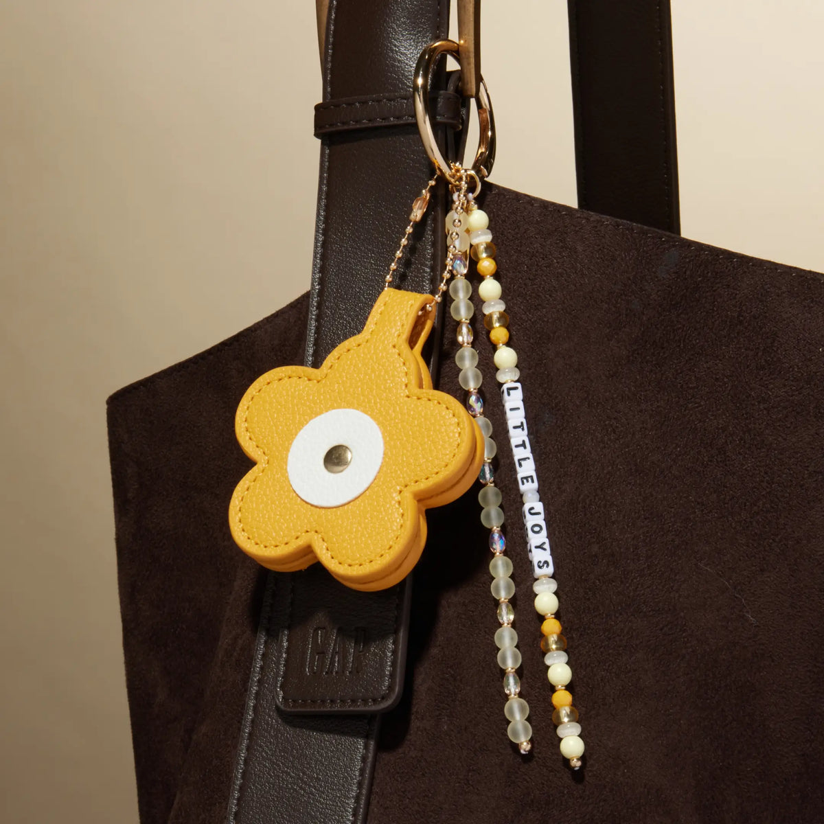Little Joys Bag Charm by Anna Sitar clipped to a brown suede tote, mustard yellow faux leather flower with the Little Joys word strand and pearl and yellow beaded tassels, lifestyle shot