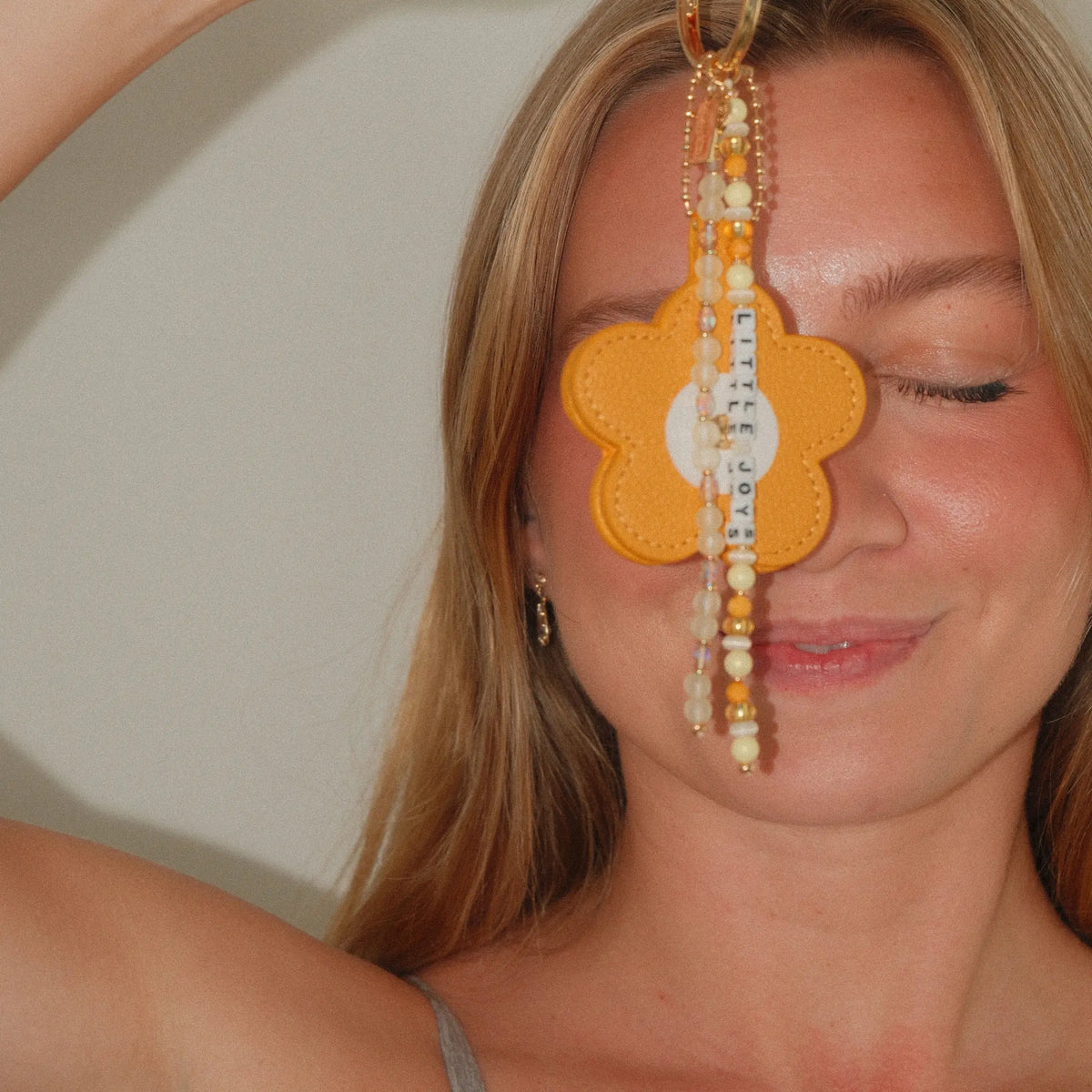 Model holding the Little Joys Bag Charm by Anna Sitar up to her face, mustard yellow faux leather flower with pearl and yellow beaded tassels on a gold key ring, on-model