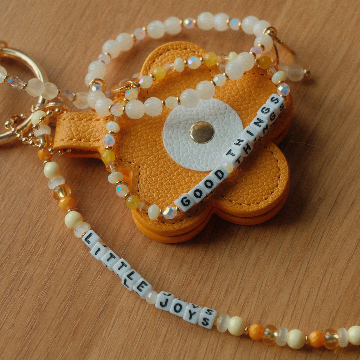 Little Joys Bag Charm by Anna Sitar with the Good Things bracelet on a wood surface, mustard yellow faux leather flower, Little Joys word strand, and yellow and white beads, detail