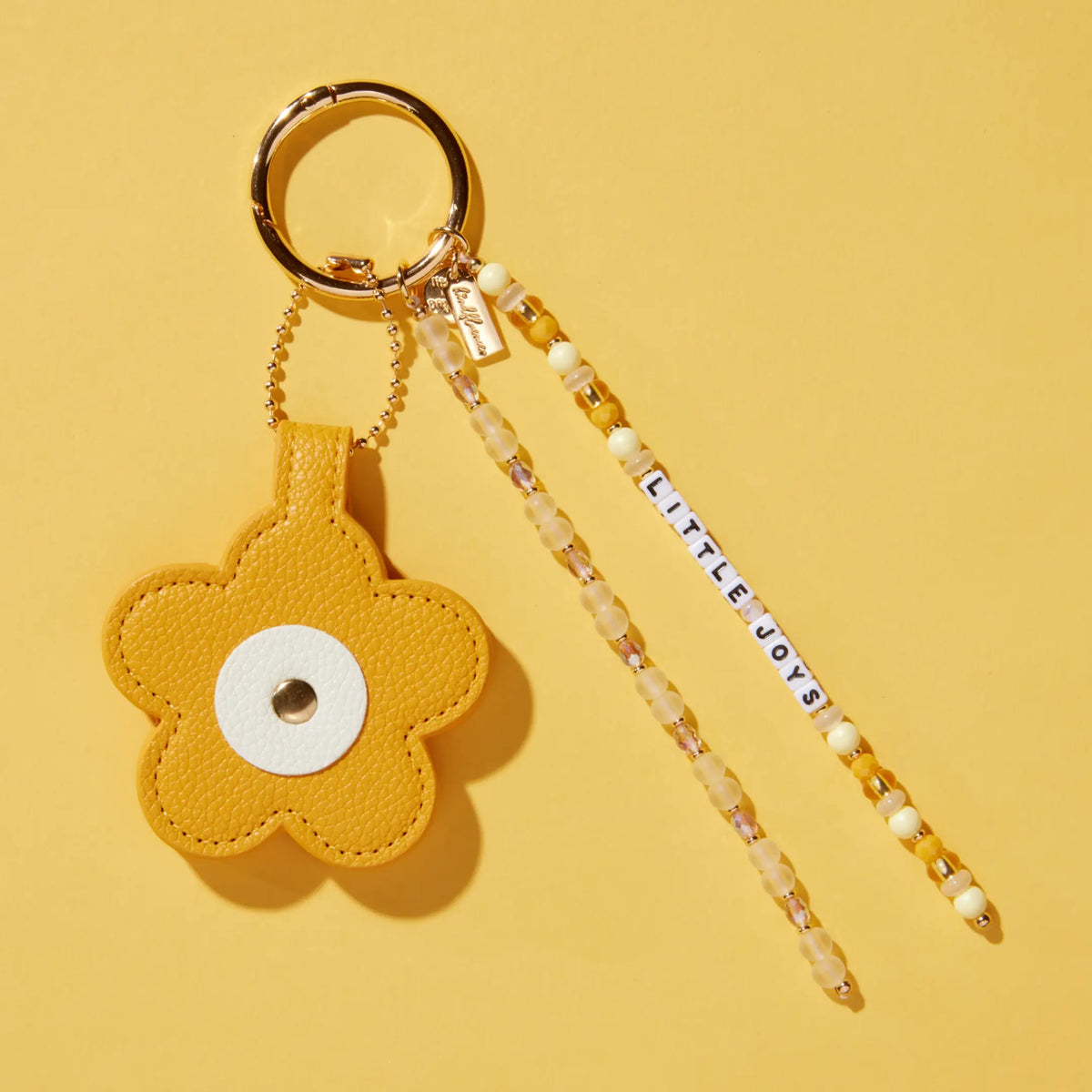 Little Joys Bag Charm by Anna Sitar, mustard yellow faux leather flower with a white center, Little Joys word strand, and beaded tassels on a gold key ring, flat-lay on yellow background