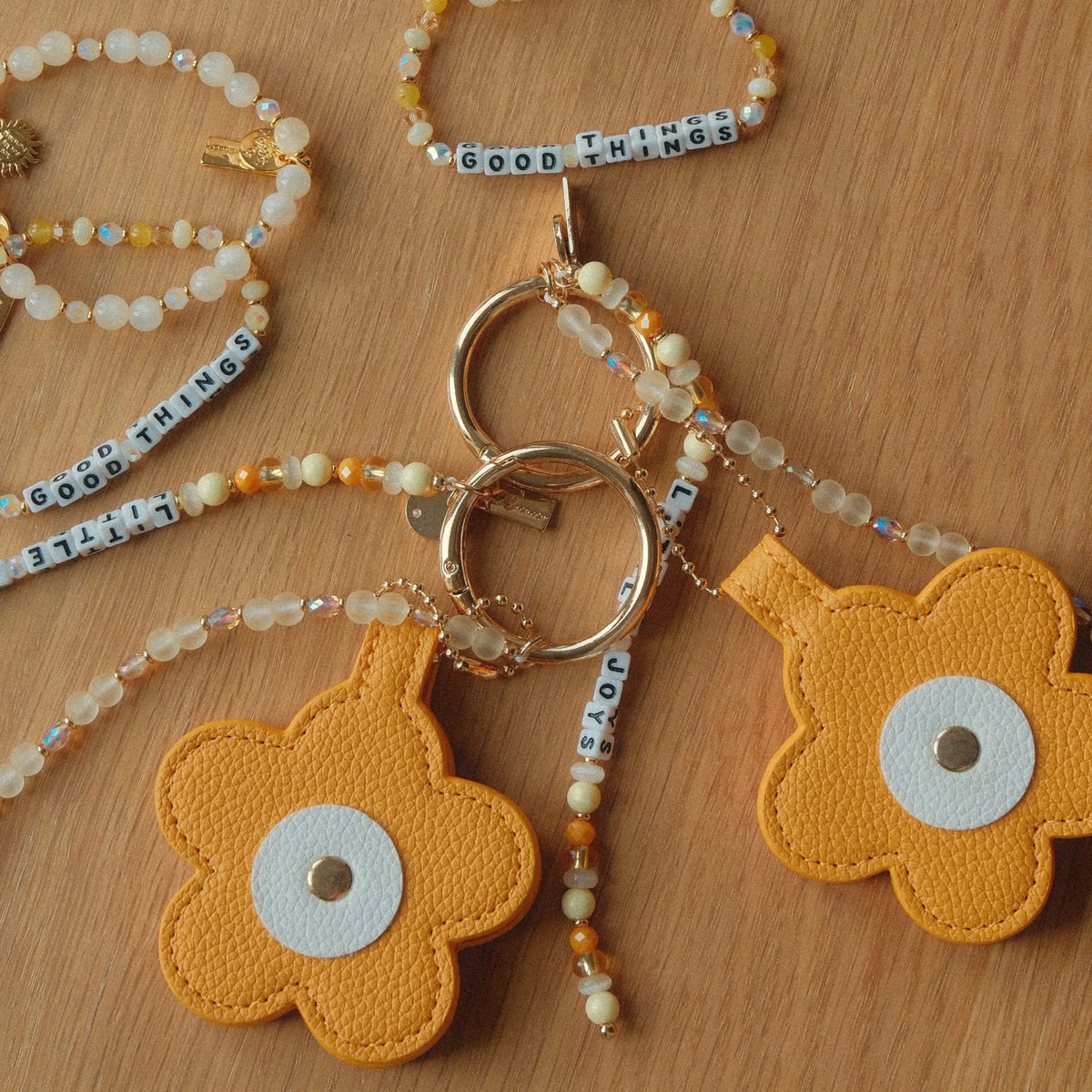 Good Things bracelets and Little Joys Bag Charms by Anna Sitar on a wood surface, white letter beads with yellow and white beads and mustard yellow faux leather flowers on gold key rings, flat-lay