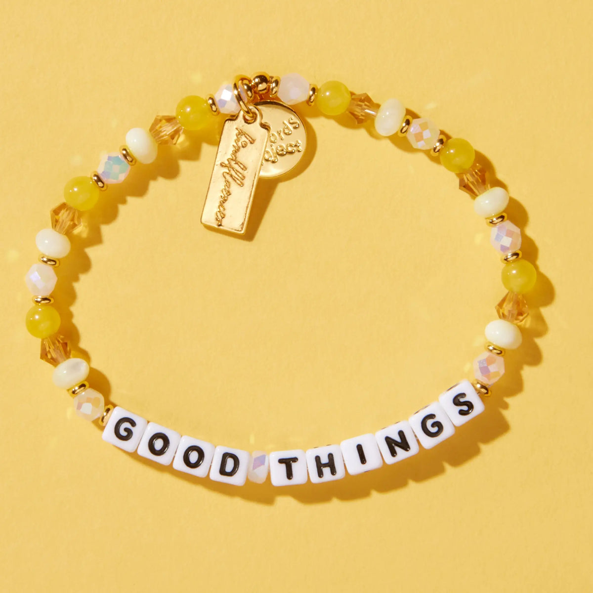Good Things bracelet by Anna Sitar in the Honeycomb bead pattern, white letter beads with yellow glass, cream, and iridescent crystal beads and a gold Kindfluencer tag, flat-lay on yellow background