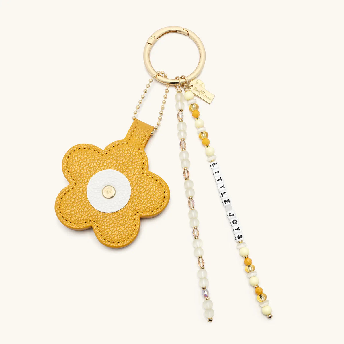 Little Joys Bag Charm by Anna Sitar, mustard yellow faux leather flower with a white center, Little Joys word strand, and pearl and yellow beaded tassels on a gold key ring, flat-lay on cream background