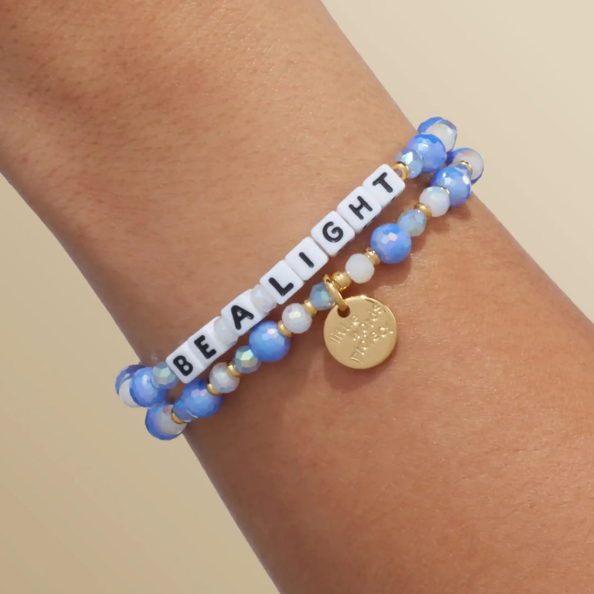Be a Light bracelet in the Blizzard Baby bead pattern with white letter beads, stacked with a second bracelet, on-model wrist shot on warm neutral background