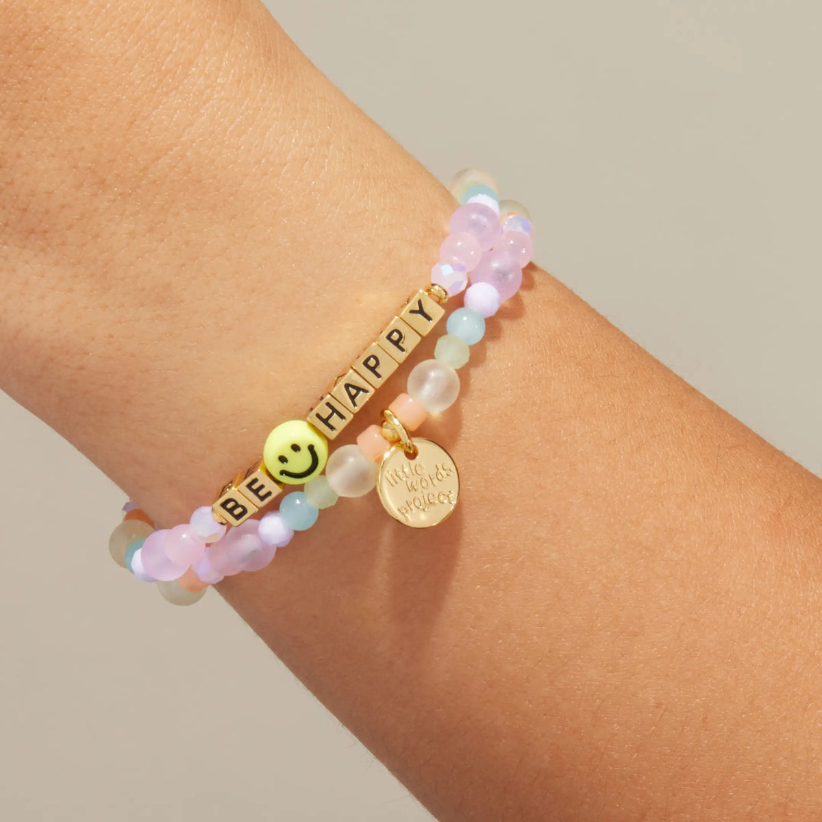 Gold Be Happy bracelet in the Radiant Bliss bead pattern with gold letter beads, stacked with a coordinating beaded bracelet, on-model on wrist against cream background
