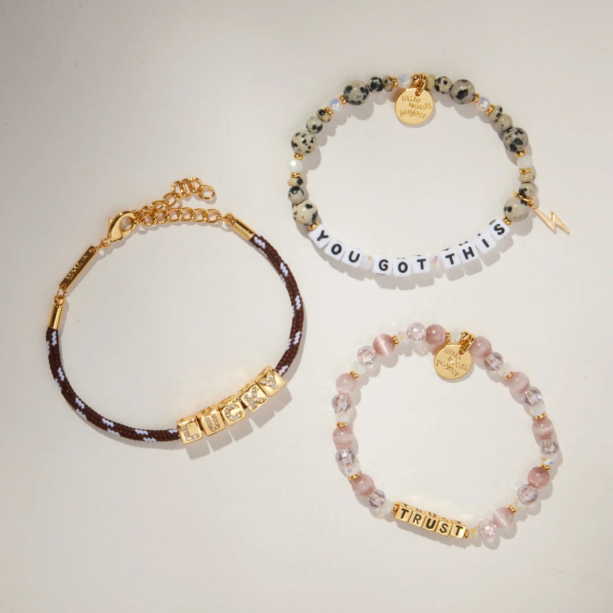Lucky Charm Set flat-lay on cream background showing three stacked bracelets: BOLDER Lucky paracord bracelet, Trust bracelet in gold letter beads, and You Got This dalmatian jasper bracelet