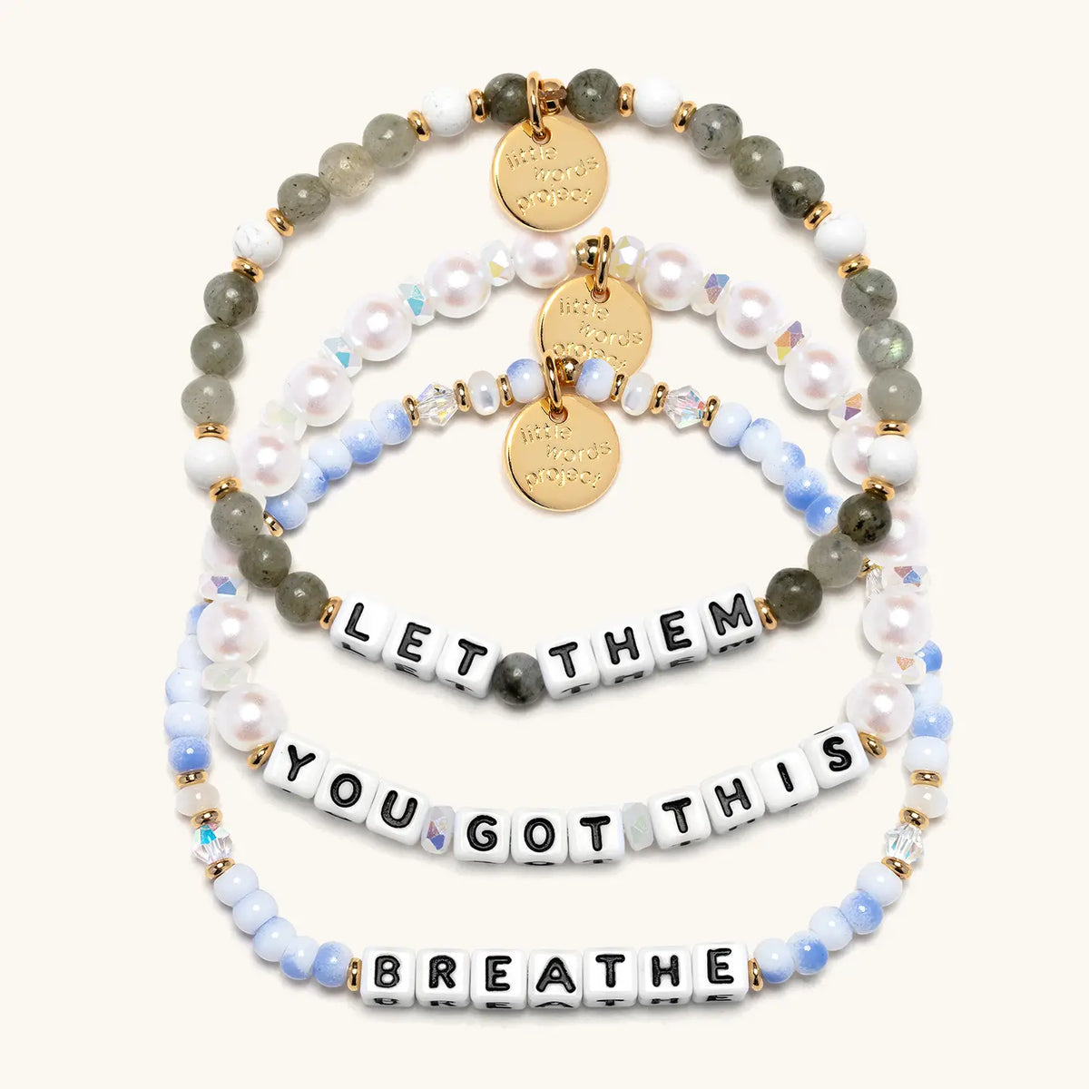 The Reset Bracelet Set in white letter beads, flat-lay on cream background showing three stacked bracelets: Let Them in gray and white stone beads, You Got This in white faux pearls and iridescent crystals, and Breathe in blue and white swirl beads
