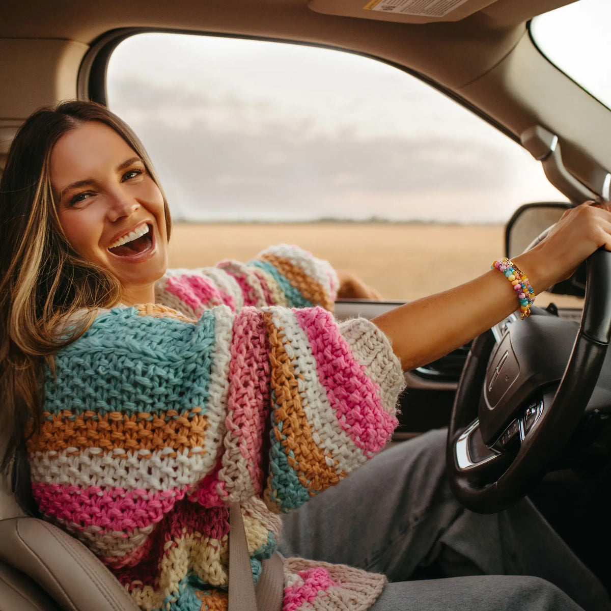 Woman laughing in the driver's seat in a pastel striped chunky knit cardigan, wearing the LWP x Britt Horton Fearless bracelet stack with gold letter beads and rainbow semi precious stone beads