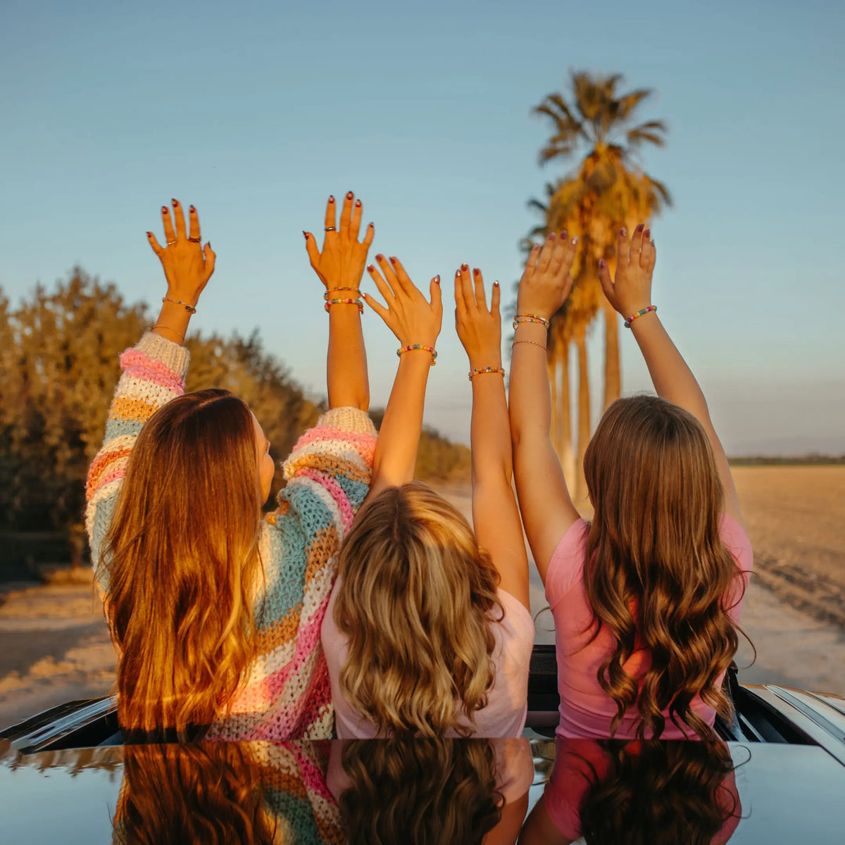 Three friends standing through a car sunroof with arms raised toward palm trees at sunset, wearing LWP x Britt Horton beaded bracelets in rainbow pastel beads