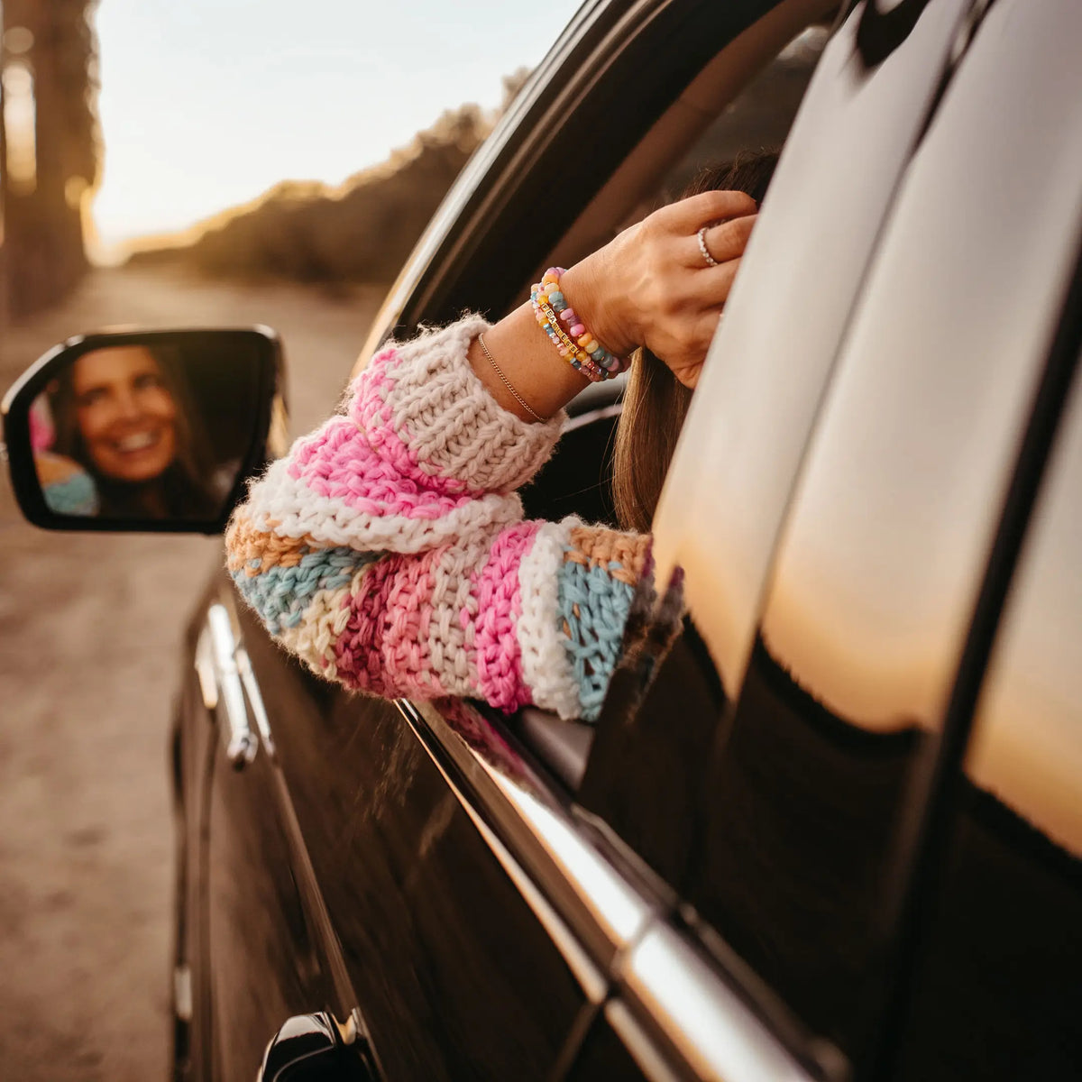 Arm resting out a car window at golden hour in a pink, blue, and cream knit sleeve, wearing the LWP x Britt Horton Fearless bracelet in gold letter beads stacked with pastel beaded bracelets