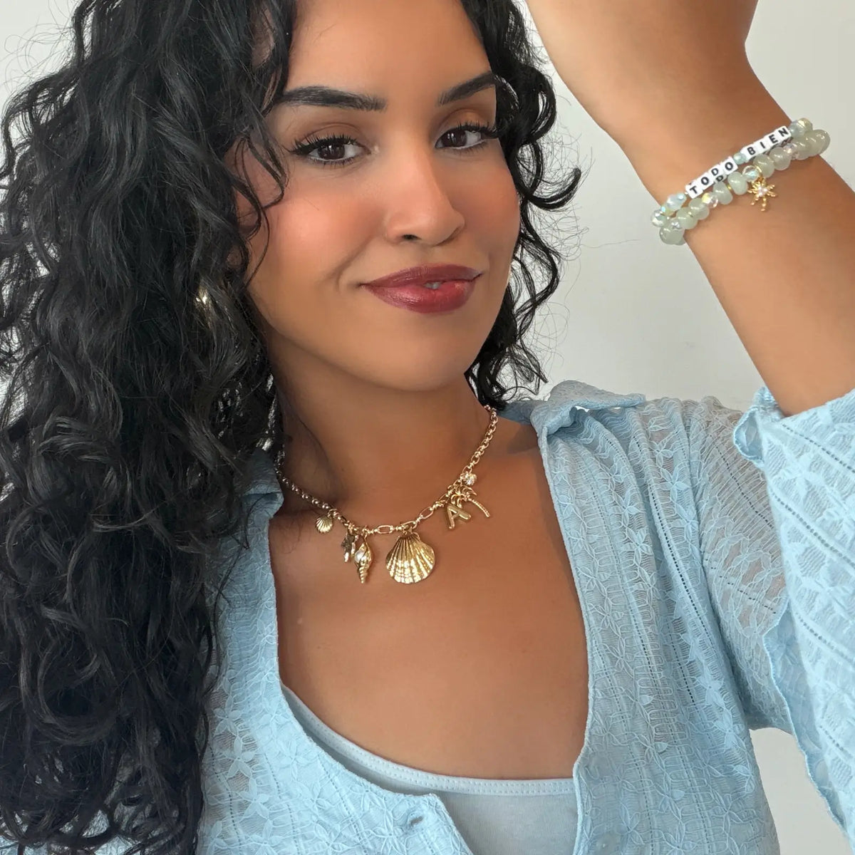 Alexia Del Valle wearing the Todo Bien Bracelet Set stacked on her wrist with a gold seashell charm necklace and light blue lace top