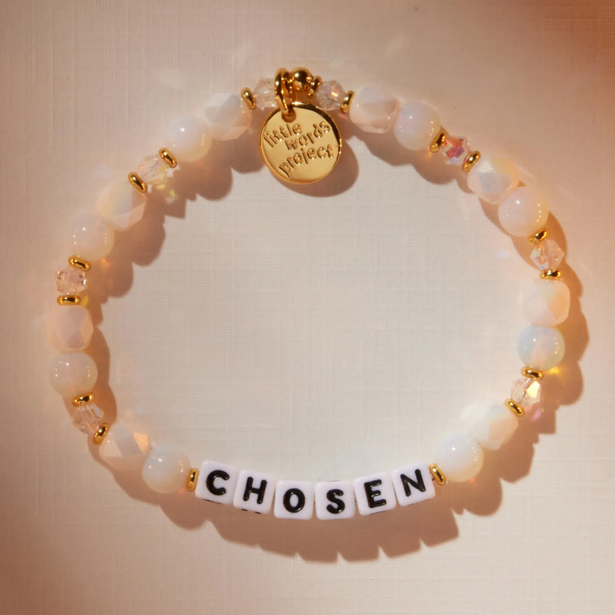 Chosen bracelet in the Snow Fantasy bead pattern with white letter beads, flat-lay on warm neutral background with soft light