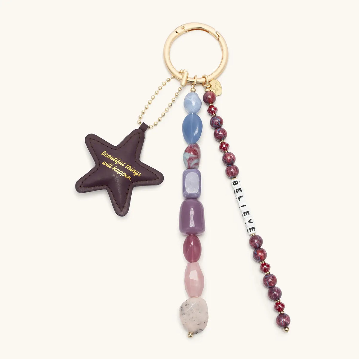 Beautiful Things Can Happen Bag Charm, plum faux leather star, beaded tassel strands in blue, purple, pink, and garnet with a Believe letter strand, on a gold round spring ring, flat-lay on cream background