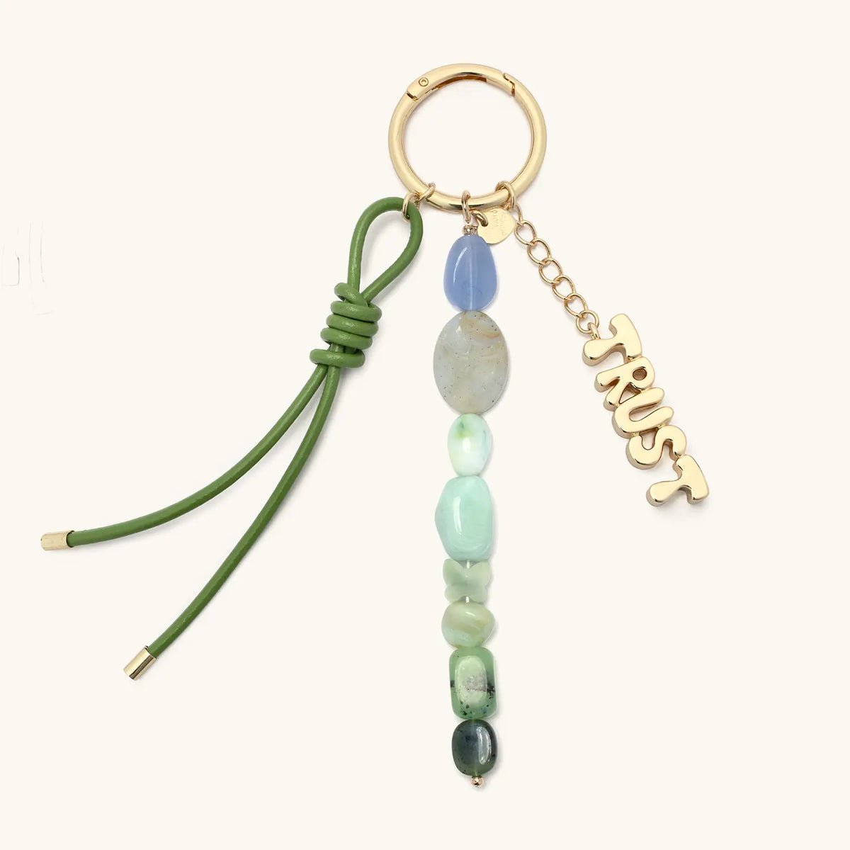 Trust Bag Charm, olive green knotted cord, beaded strand in blue, sage, and green stone beads, and gold Trust script charm on a gold chain, on a gold round spring ring, flat-lay on cream background