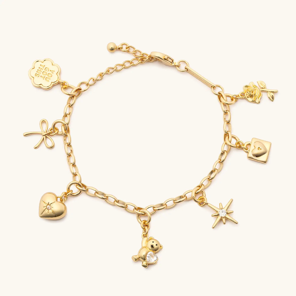 Rolo Chain Charm Bracelet, gold rolo chain bracelet with seven gold charms: You Got This coin, bow, cz heart, teddy bear gem, starburst, padlock, and rose, flat-lay on cream background