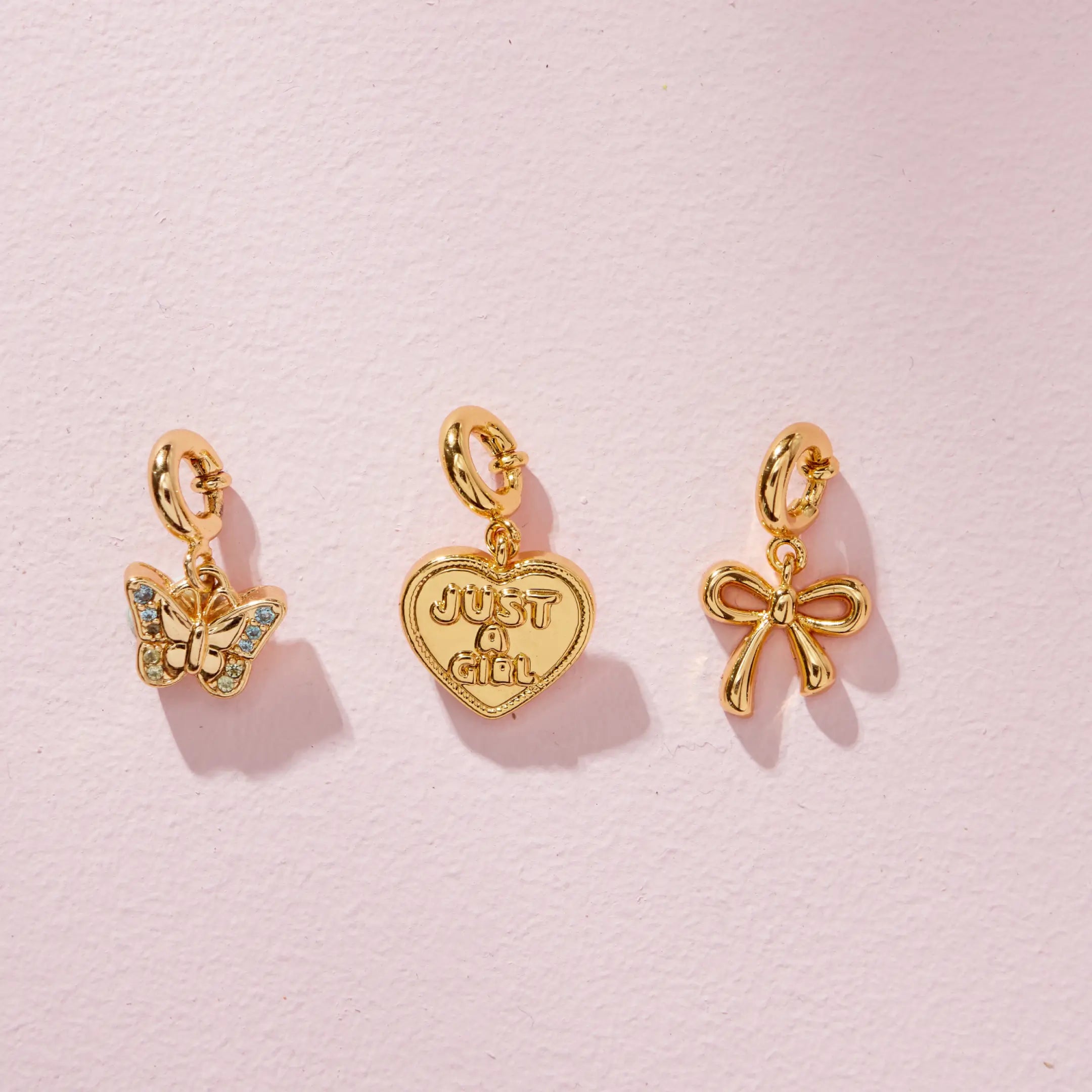 Just A Girl Charm Set trio of gold charms, flat lay on pink