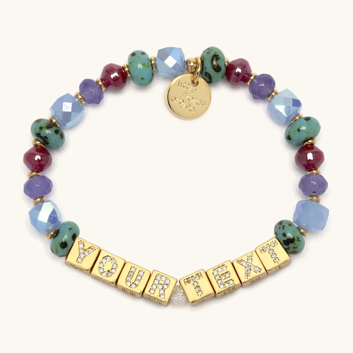 BOLDER Custom Jewel Tones bracelet in the Jewel Tones bead pattern, gold CZ letter blocks reading Your Text, teal speckled, plum, burgundy, periwinkle, and light blue faceted beads with gold accents, flat-lay on cream background