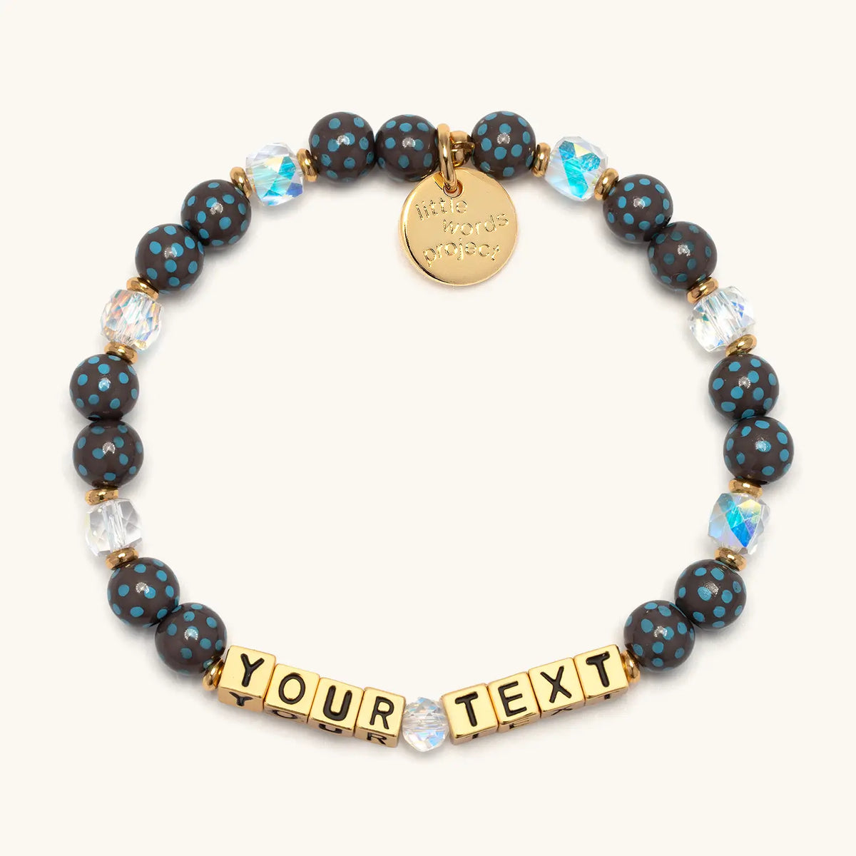 Custom Polky Babe bracelet with gold letter beads reading Your Text, dark gray beads with blue polka dots and clear iridescent crystals with gold accents, flat-lay on cream background