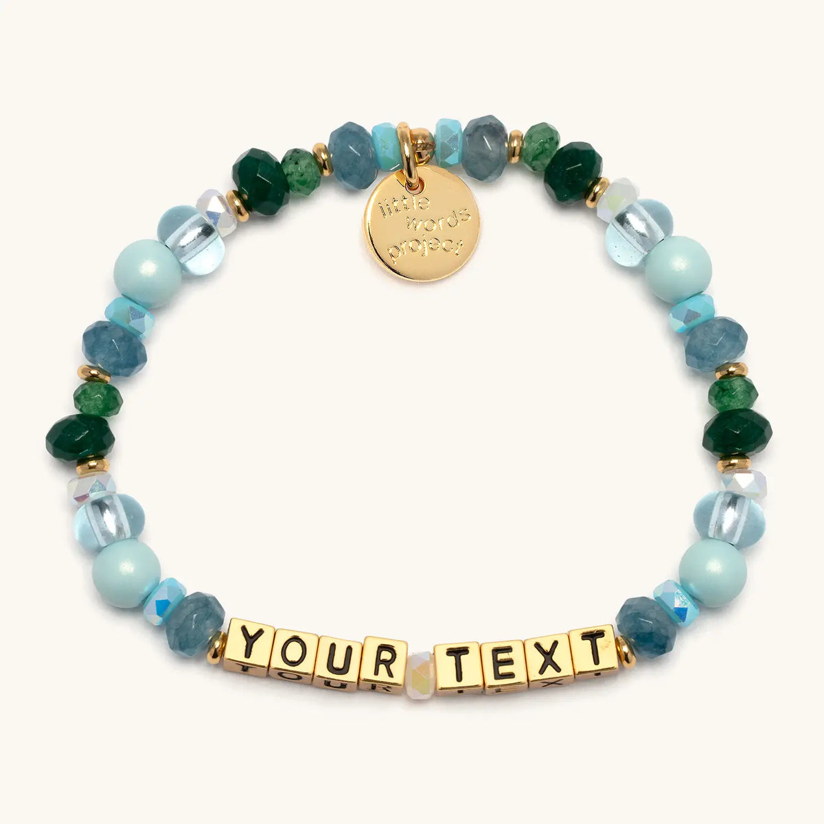 Custom Rebel Cozy bracelet with gold letter beads reading Your Text, light blue pearls, teal faceted beads, emerald green crystals, and clear glass beads with gold accents, flat-lay on cream background