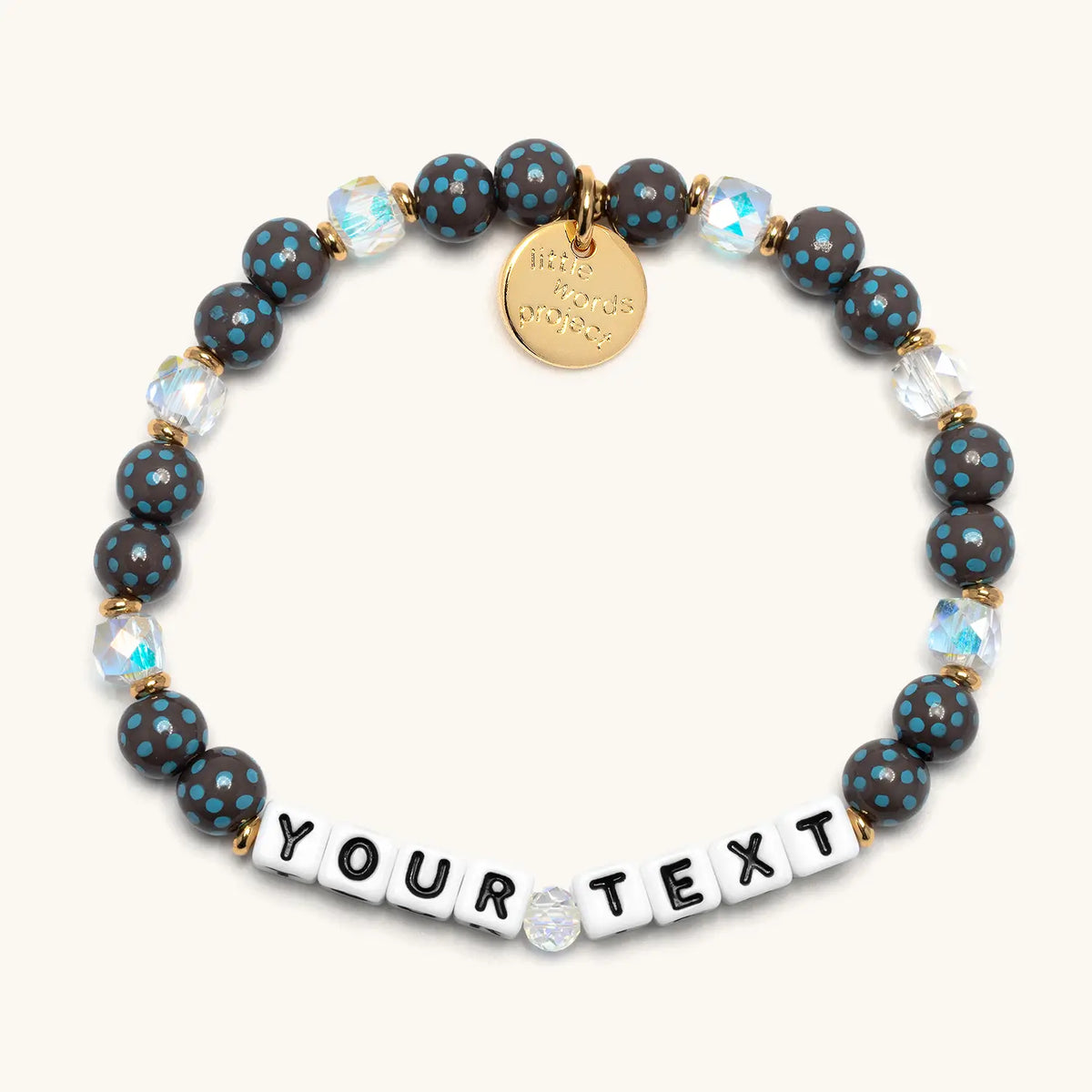Custom Polky Babe bracelet with white letter beads reading Your Text, dark gray beads with blue polka dots and clear iridescent crystals with gold accents, flat-lay on cream background