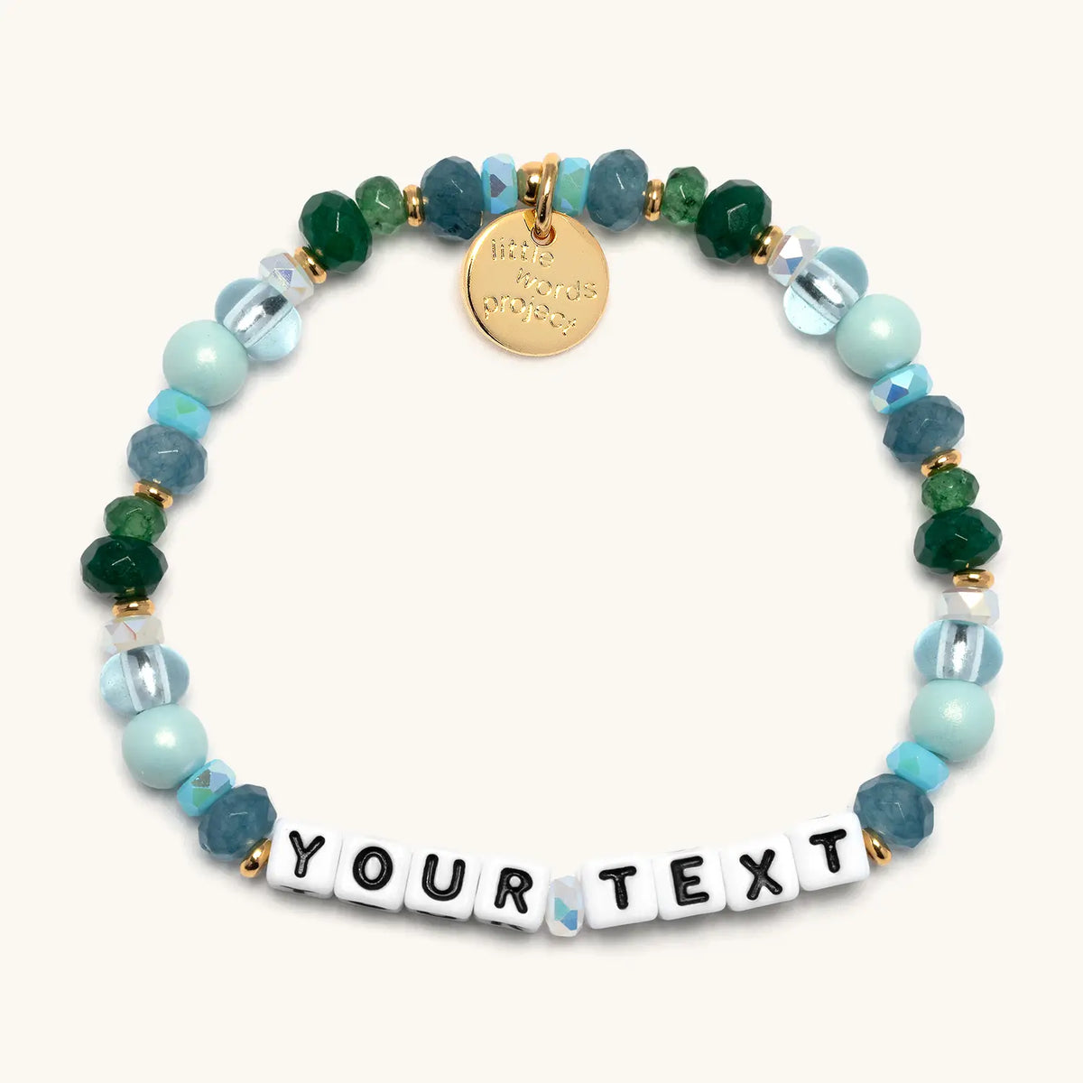 Custom Rebel Cozy bracelet with white letter beads reading Your Text, light blue pearls, teal faceted beads, emerald green crystals, and clear glass beads with gold accents, flat-lay on cream background