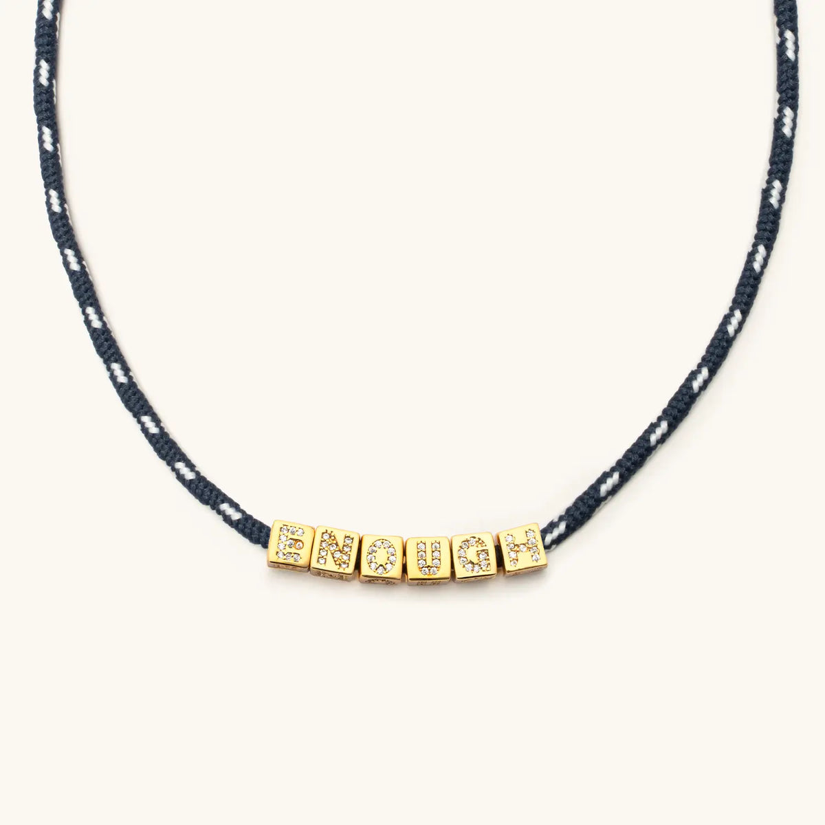 Close-up of BOLDER Enough Necklace, navy and white flecked paracord with gold CZ letter blocks spelling Enough, flat-lay on cream background