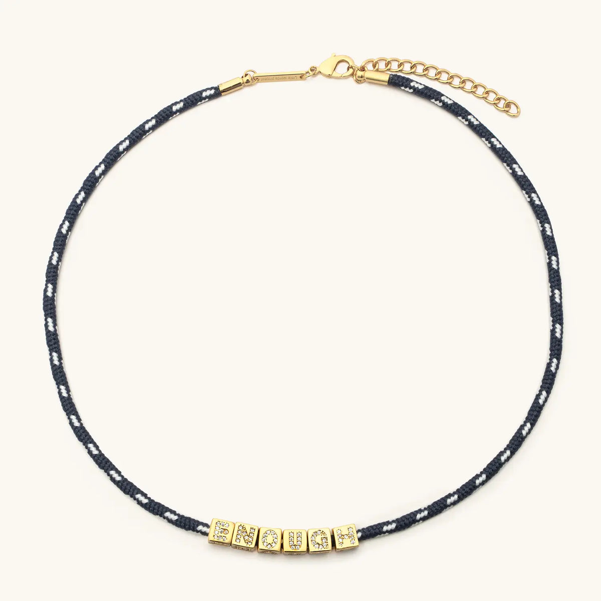 BOLDER Enough Necklace, navy and white flecked paracord with gold CZ letter blocks, gold bar ends, lobster clasp, and chain extender, flat-lay on cream background