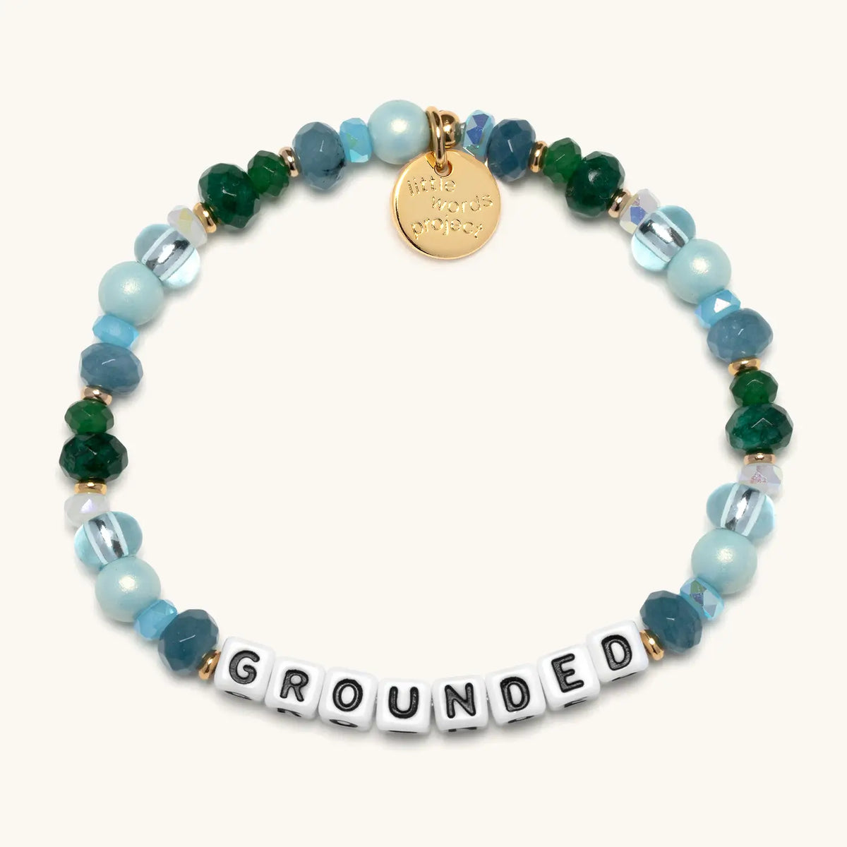 Grounded bracelet in the Rebel Cozy bead pattern, white letter beads with light blue pearls, teal faceted beads, emerald green crystals, and clear glass beads with gold accents, flat-lay on cream background