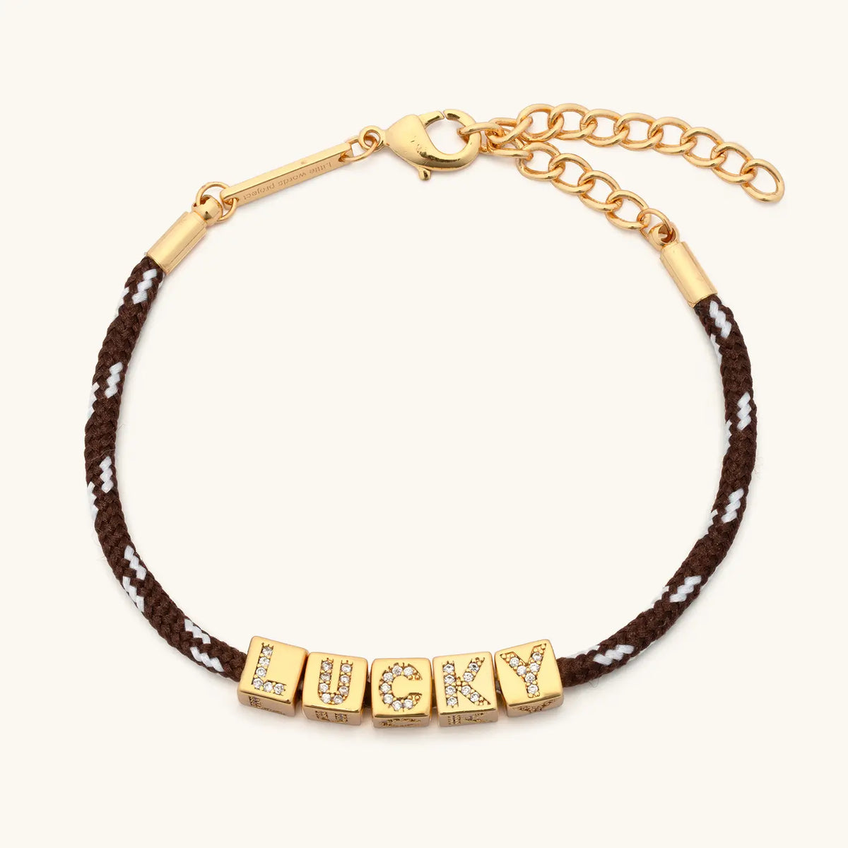BOLDER Lucky Bracelet, brown and white flecked paracord with gold CZ letter blocks, gold bar ends, lobster clasp, and chain extender, flat-lay on cream background