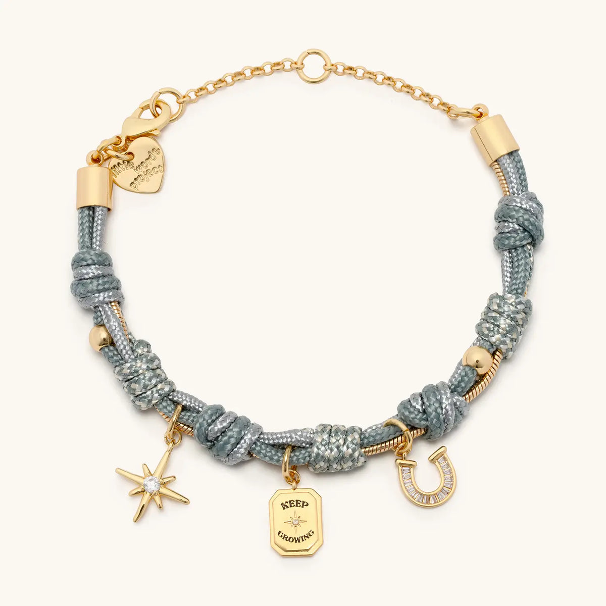 Keep Growing Paracord Charm Bracelet, sage gray knotted paracord with gold chain extender and three gold charms: a starburst, a Keep Growing tag, and a CZ horseshoe, flat-lay on cream background