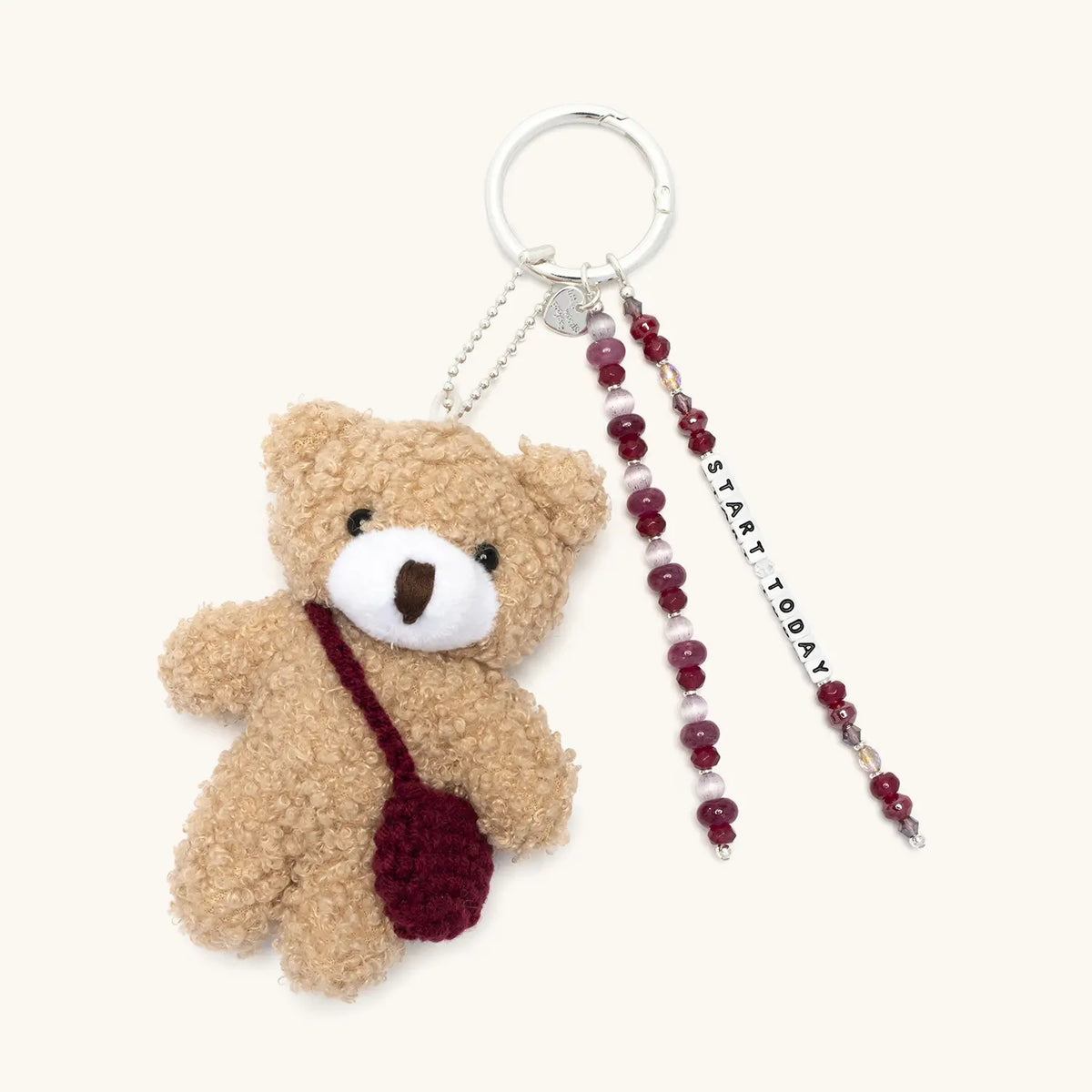Start Today Bag Charm, tan plush teddy bear with a burgundy crochet crossbody bag, burgundy and pearl beaded Start Today word strand, and beaded strand on a silver key ring, flat-lay on cream background