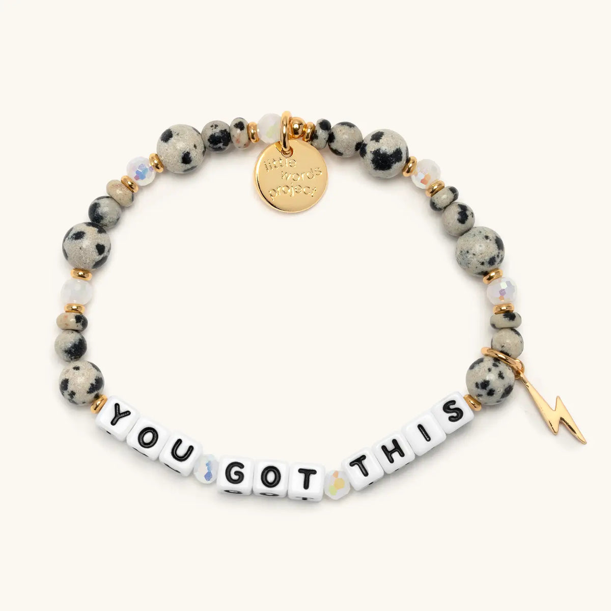 You Got This bracelet in the Dot Vibes bead pattern, white letter beads with dalmatian jasper stone beads, iridescent crystals, gold accents, and a gold lightning bolt charm, flat-lay on cream background