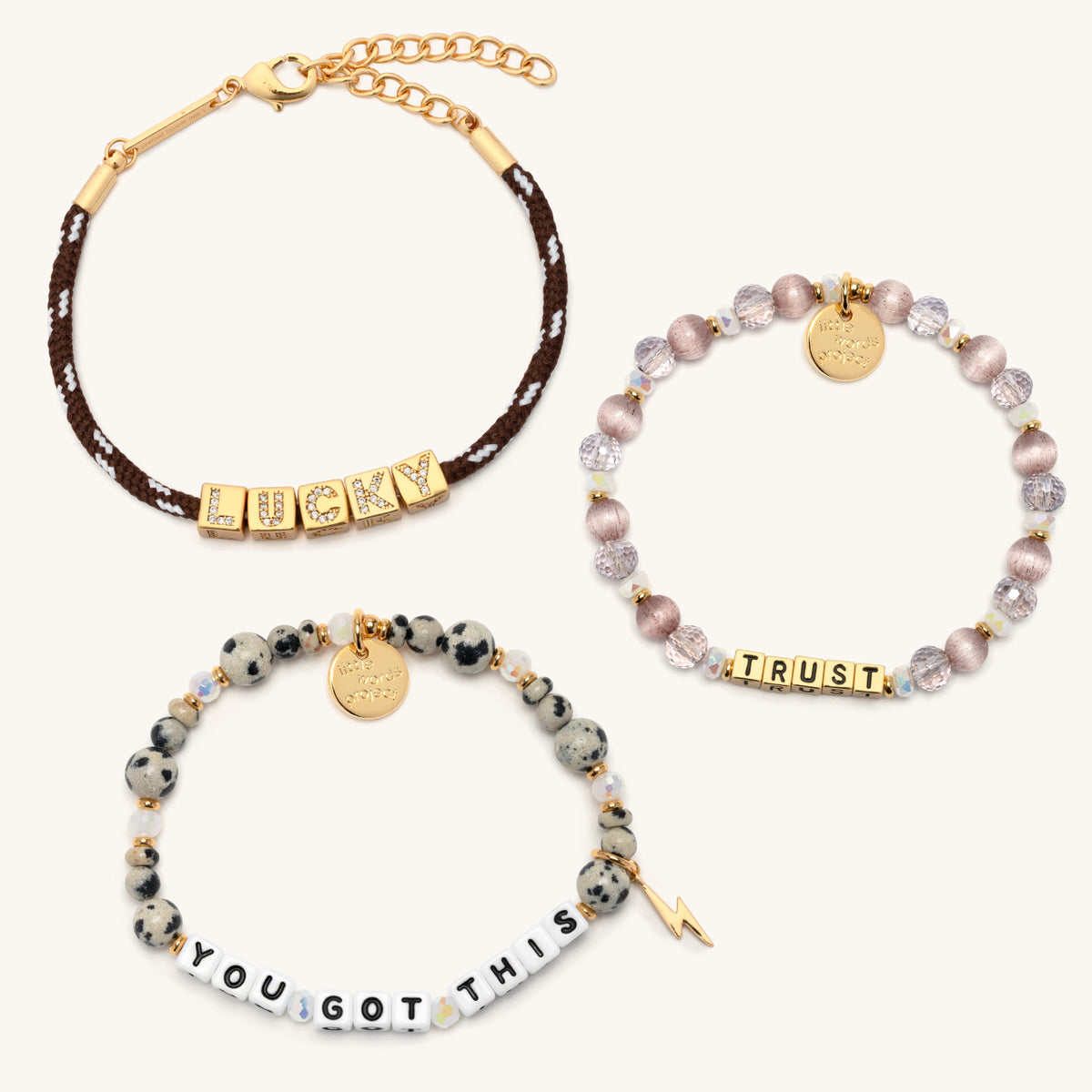 Lucky Charm Set bracelets laid out separately on cream background: BOLDER Lucky brown paracord bracelet, Trust bracelet with gold letter beads and mauve crystal beads, and You Got This dalmatian jasper bracelet with gold lightning bolt charm