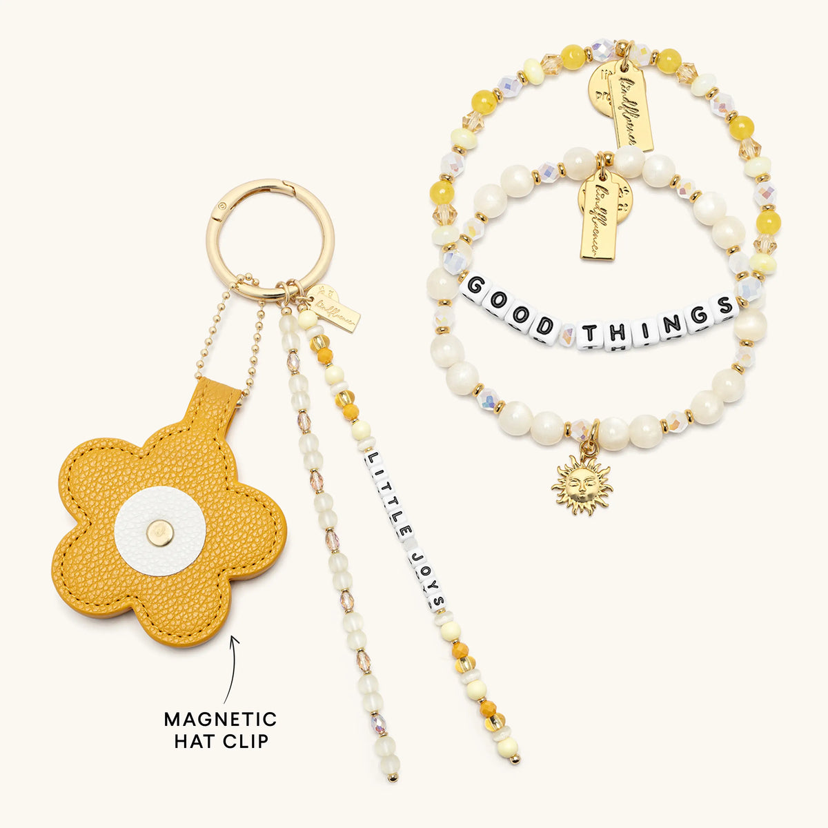 A coordinated jewelry and accessory set on a light cream background: a mustard yellow flower-shaped magnetic hat clip with a white center, a beaded bracelet with pearl-like white beads, yellow beads, clear faceted beads, silver letter beads spelling GOOD THINGS, a small gold sun charm, and two gold name tags. Text on the image reads MAGNETIC HAT CLIP and LITTLE JOY.