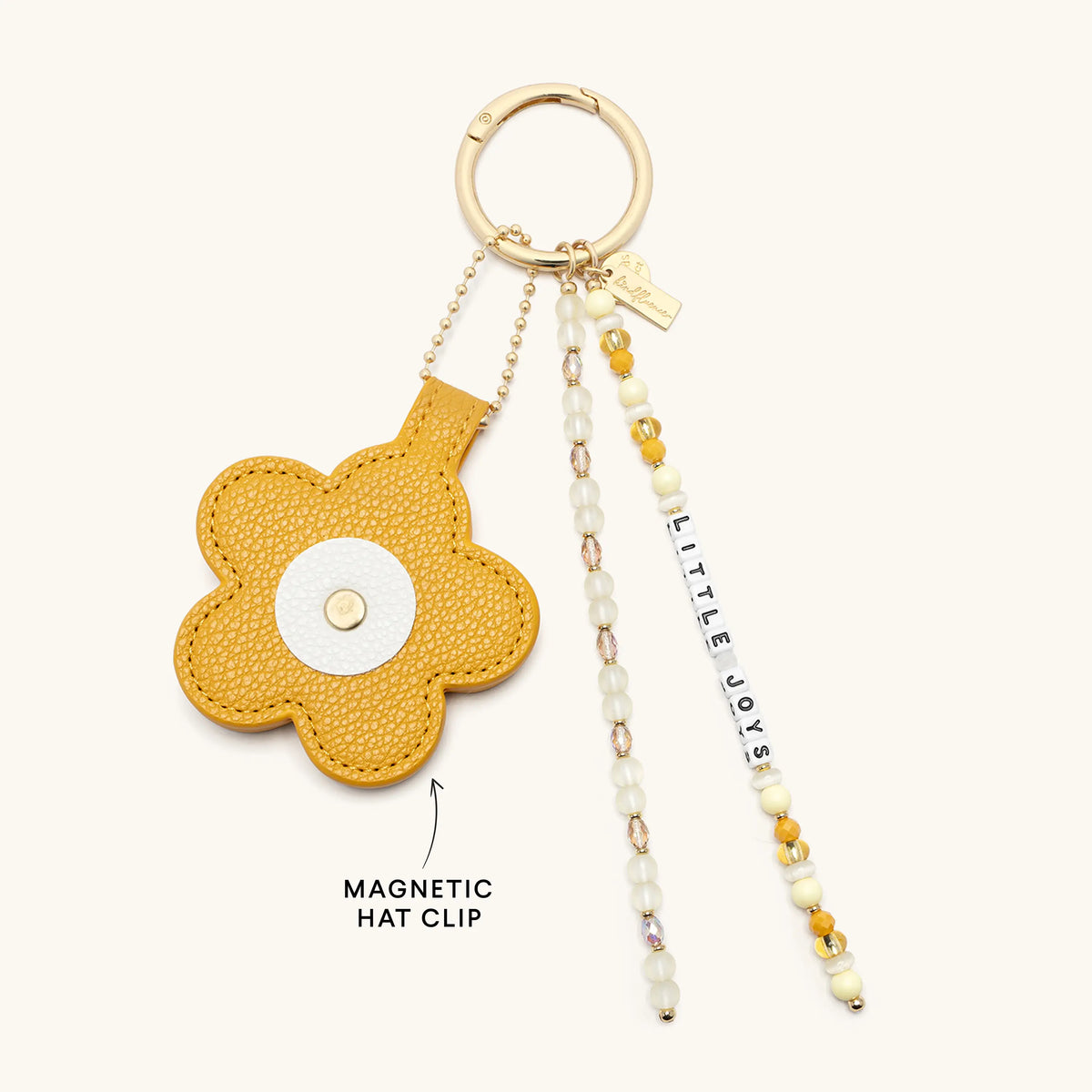 A yellow textured faux-leather flower-shaped hat clip keychain with a gold-tone ring, hanging charm beads, and a dangling tag that reads 'LITTLE JOY'. A black arrow points to the flower clip with the text 'MAGNETIC HAT CLIP' below it, all on a light cream background.