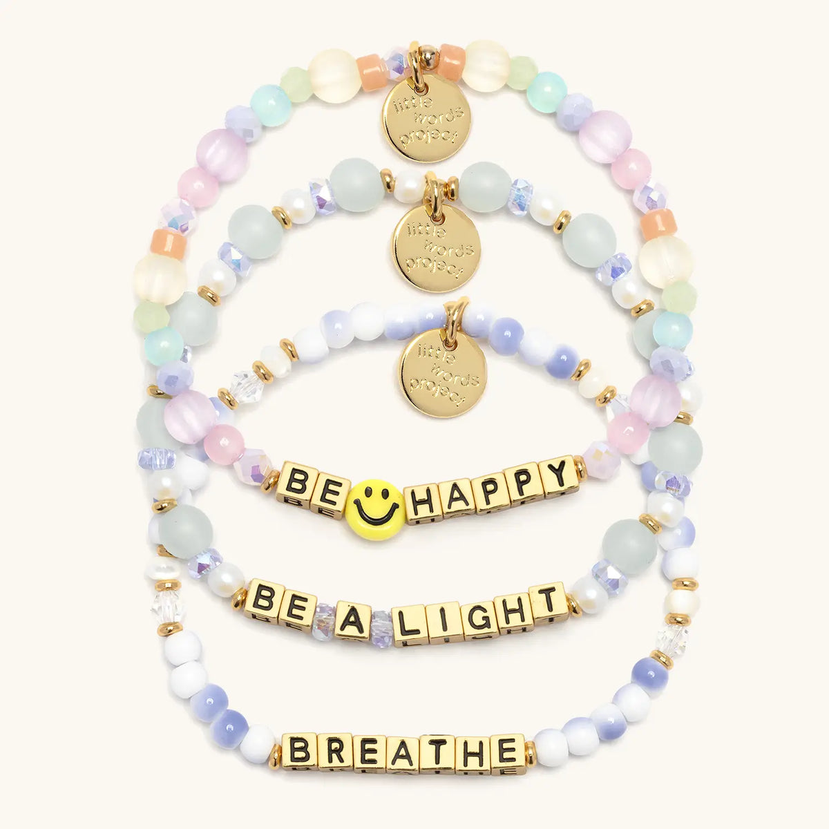 Good as Gold Bracelet Set in gold letter beads, flat-lay on cream background showing three stacked bracelets: Be Happy with a yellow smiley face bead and pastel beads, Be a Light in seafoam glass and pearl beads, and Breathe in blue and white swirl beads