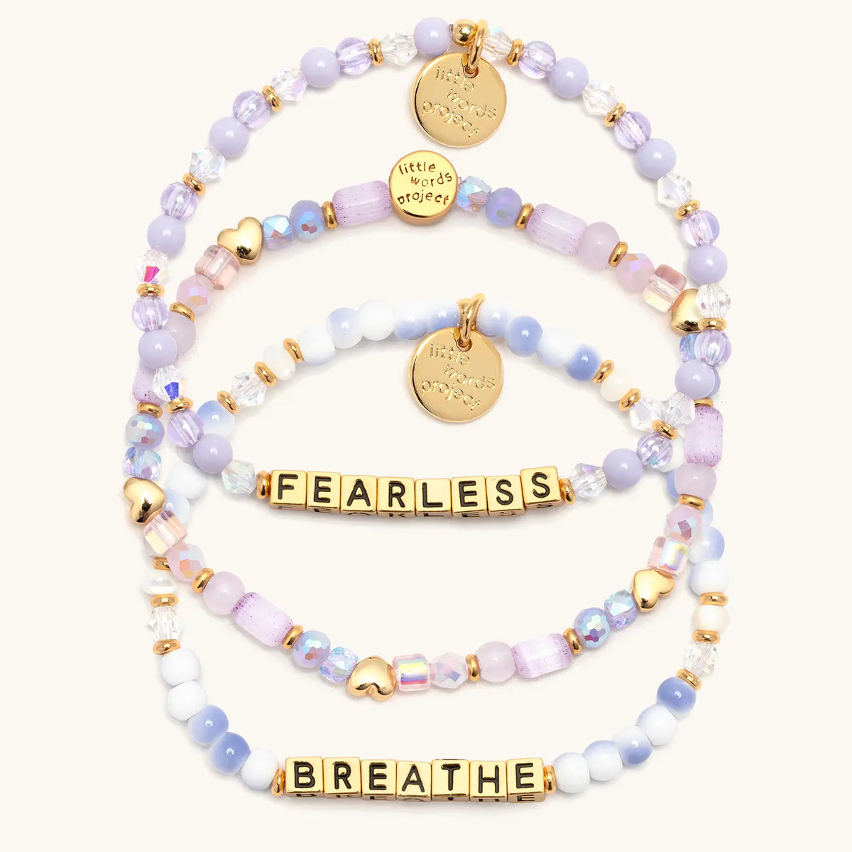 Soft & Strong Bracelet Set in gold letter beads, alternate flat-lay on cream background showing the Hearts by the Yard stacker with gold heart beads, Fearless in lavender beads and crystals, and Breathe in blue and white swirl beads