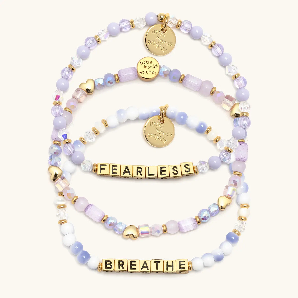 Soft & Strong Bracelet Set in gold letter beads, flat-lay on cream background showing three stacked bracelets: Fearless in lavender beads and crystals, the Hearts by the Yard stacker with gold heart beads, and Breathe in blue and white swirl beads