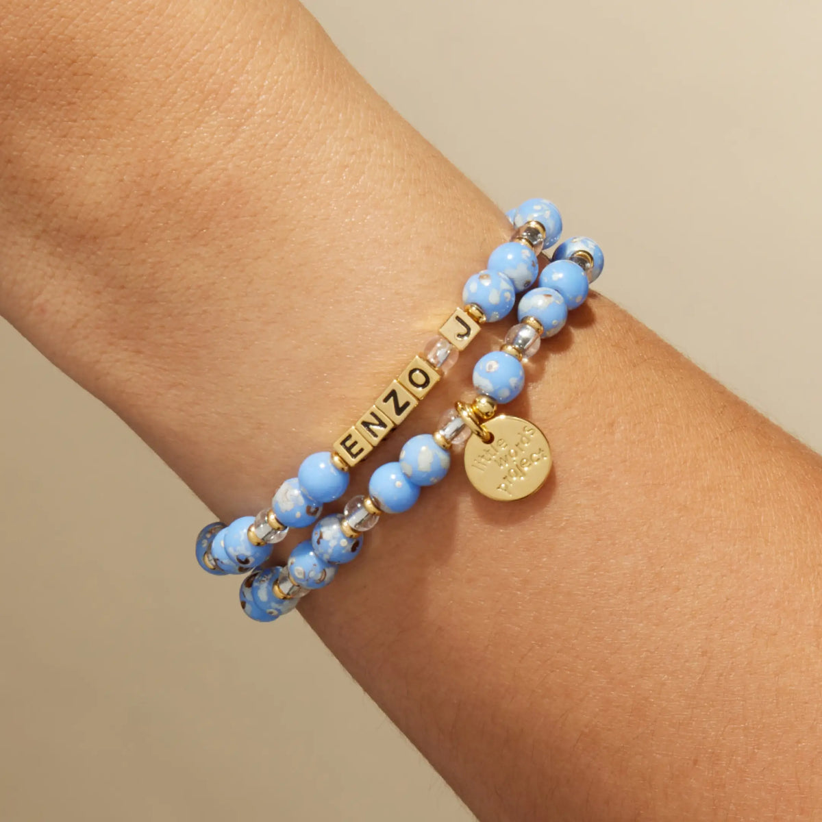 Custom Glacier Glow bracelet with gold letter beads reading Enzo, light blue speckled and clear crystal beads with a gold LWP charm, on-model on wrist against a tan background