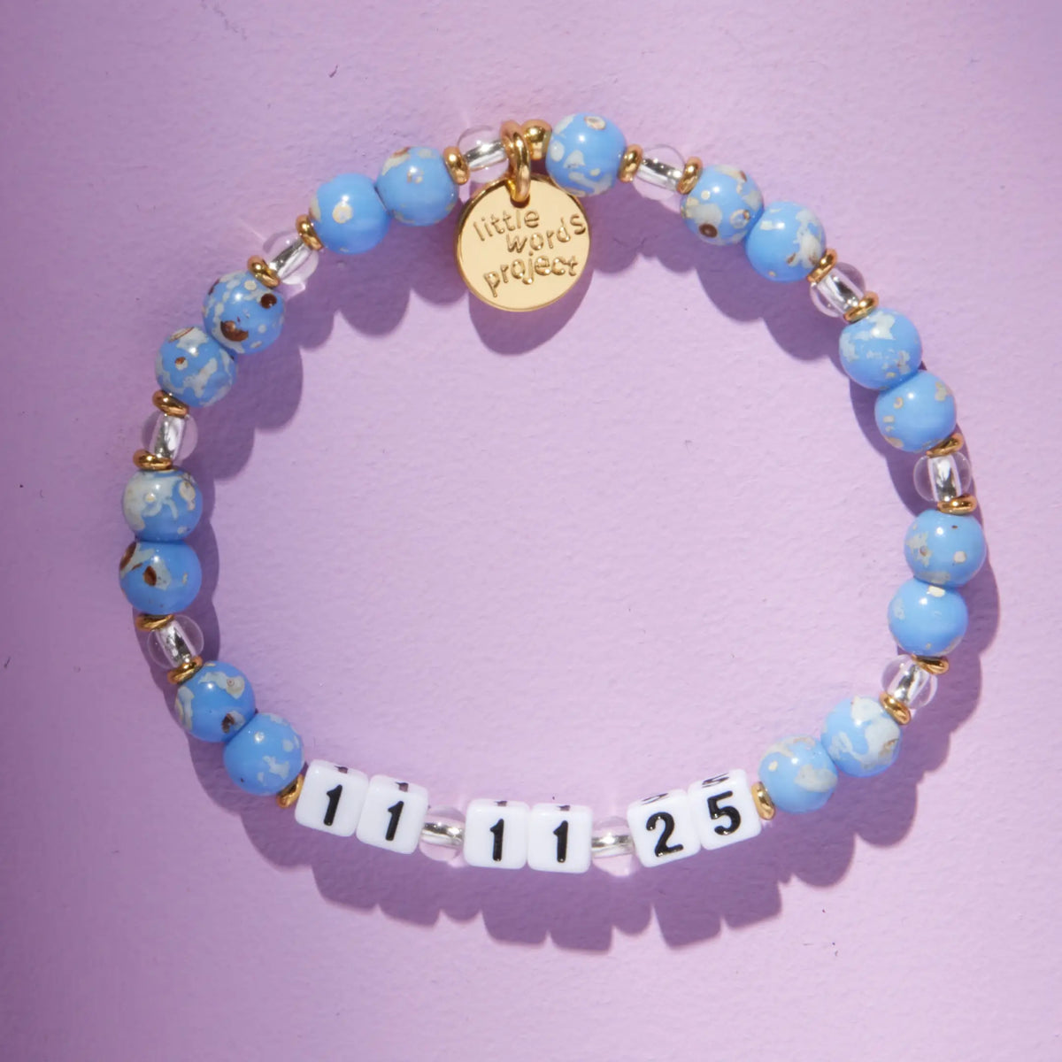 Custom Glacier Glow bracelet with white letter beads reading 11 11 25, light blue speckled and clear crystal beads with gold accents, flat-lay on lavender background